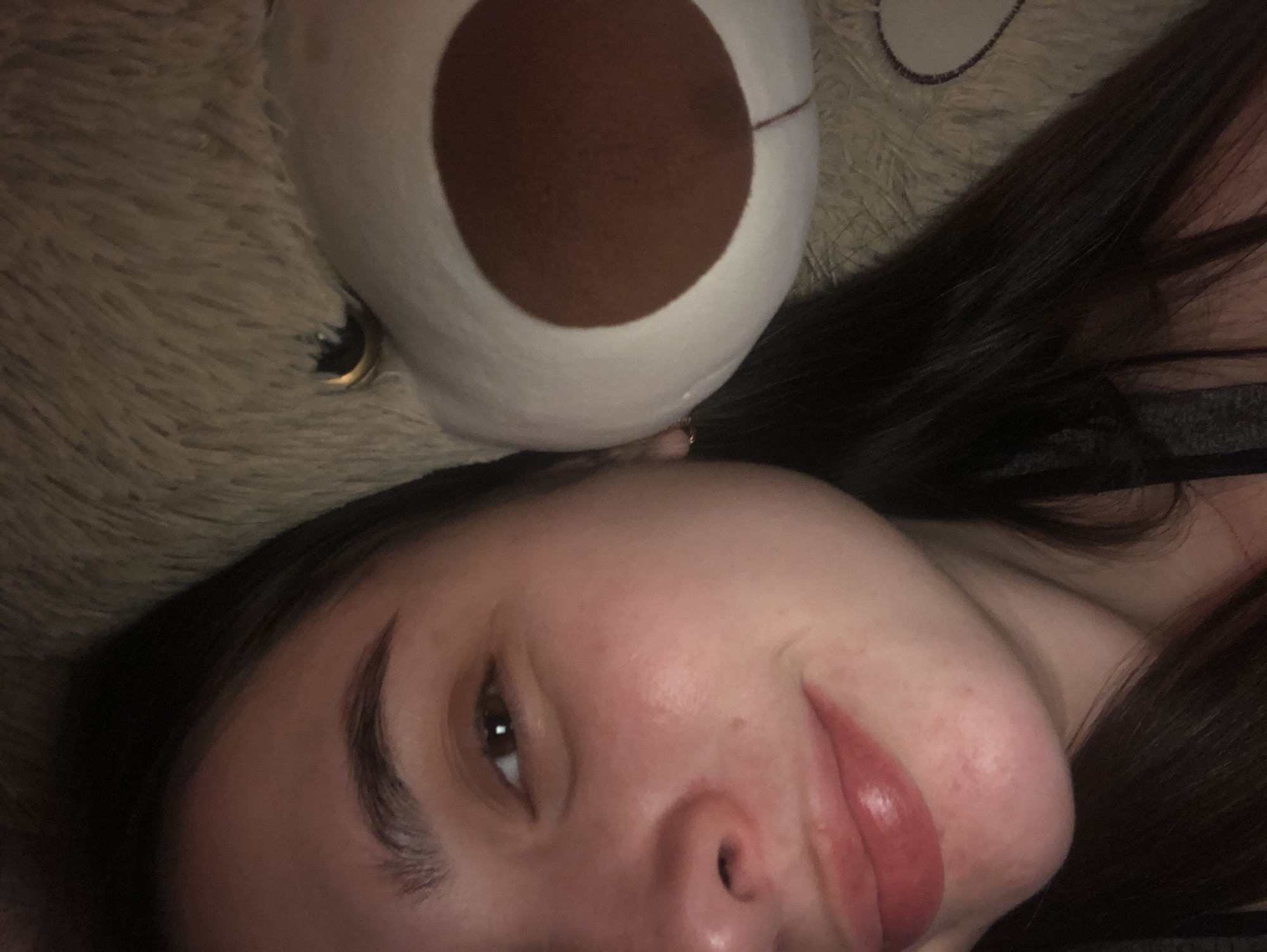 A close-up, rotated image depicts a woman lying down with a teddy bear. The woman has dark hair, fair skin, and is wearing lipstick. Part of the teddy bear's head rests near the top of the woman's head. The teddy bear is light brown with a dark brown nose. The background is blurry and out of focus. There is no indication of Noviy, Russia in this image.