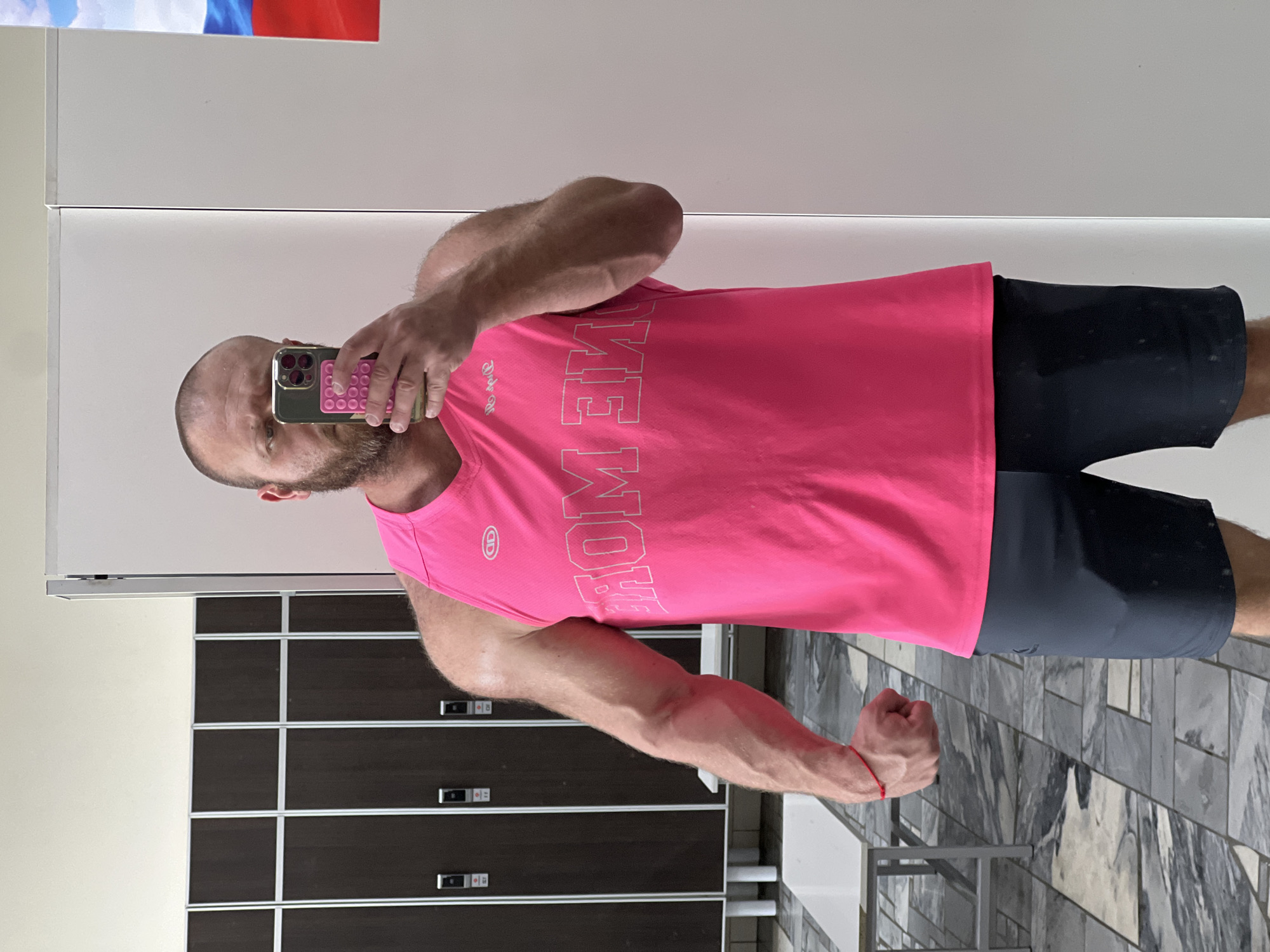 A bald, bearded man wearing a bright pink tank top with "ONE MORE" printed on it in white letters and dark grey athletic shorts is taking a selfie in a locker room. He is standing in front of a mirror, with his left arm flexed and his right hand holding a phone. The locker room has rows of dark wood lockers with silver accents, a tiled floor with a grey and white marble pattern, and a white bench. In the reflection of the mirror, a small part of the Russian flag is visible on the wall to the right. The image is set in the city of Chaikovsky, Russia.