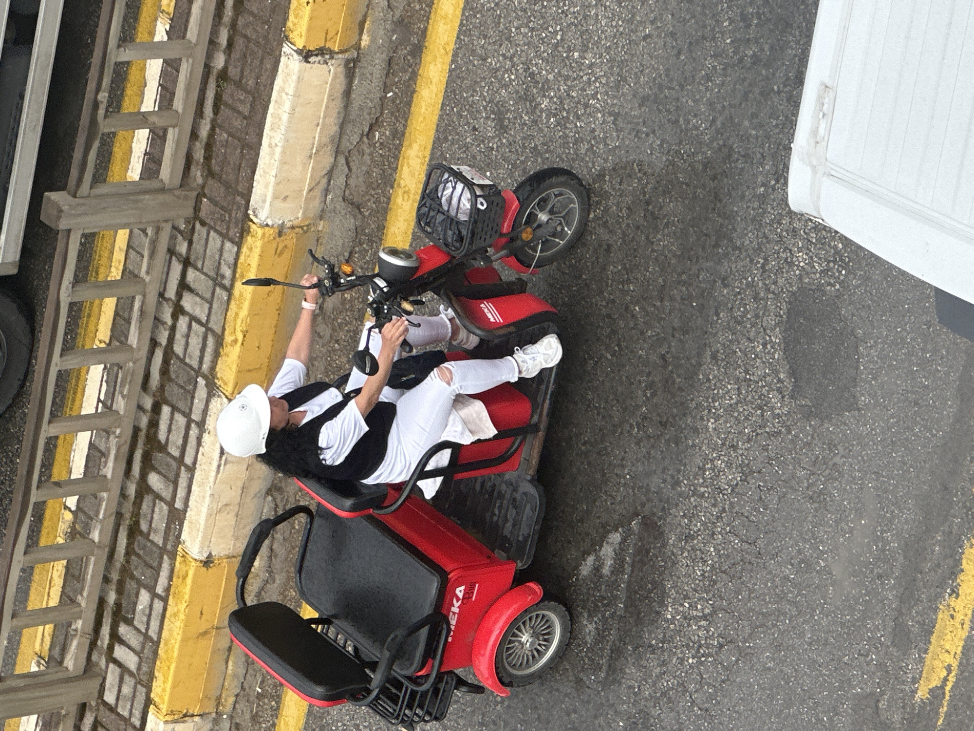 Captured from an elevated perspective, the image showcases a person seated on a red mobility scooter, positioned beside a striped curb. The individual, clad in white trousers, sneakers, a black and white top, and a white helmet, is operating the scooter. The mobility scooter is situated on asphalt. The scene suggests an urban setting in Fethiye, Turkey. A ladder is next to the striped curb. Part of a white truck is also visible.