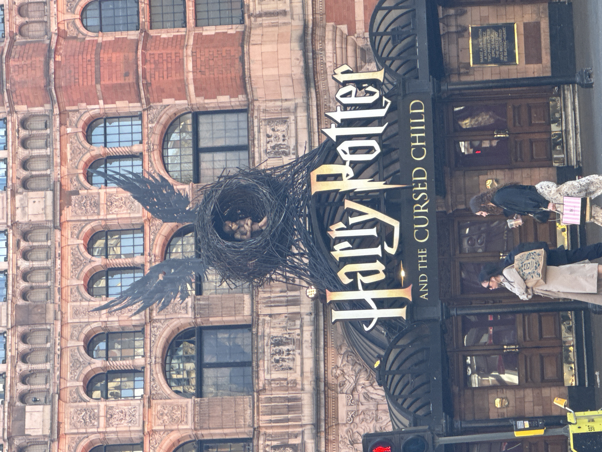 The image captures the exterior of the Palace Theatre in London, showcasing the "Harry Potter and the Cursed Child" play. A large nest with wings is centrally positioned above the theater's entrance, adorned with the play's title in bold lettering. The theater's facade features a traditional architectural style with arched windows and ornate details. People are visible walking past the entrance, including a woman carrying a shopping bag. A traffic light is visible in the foreground. A sign next to the door describes the play as a "masterpiece".