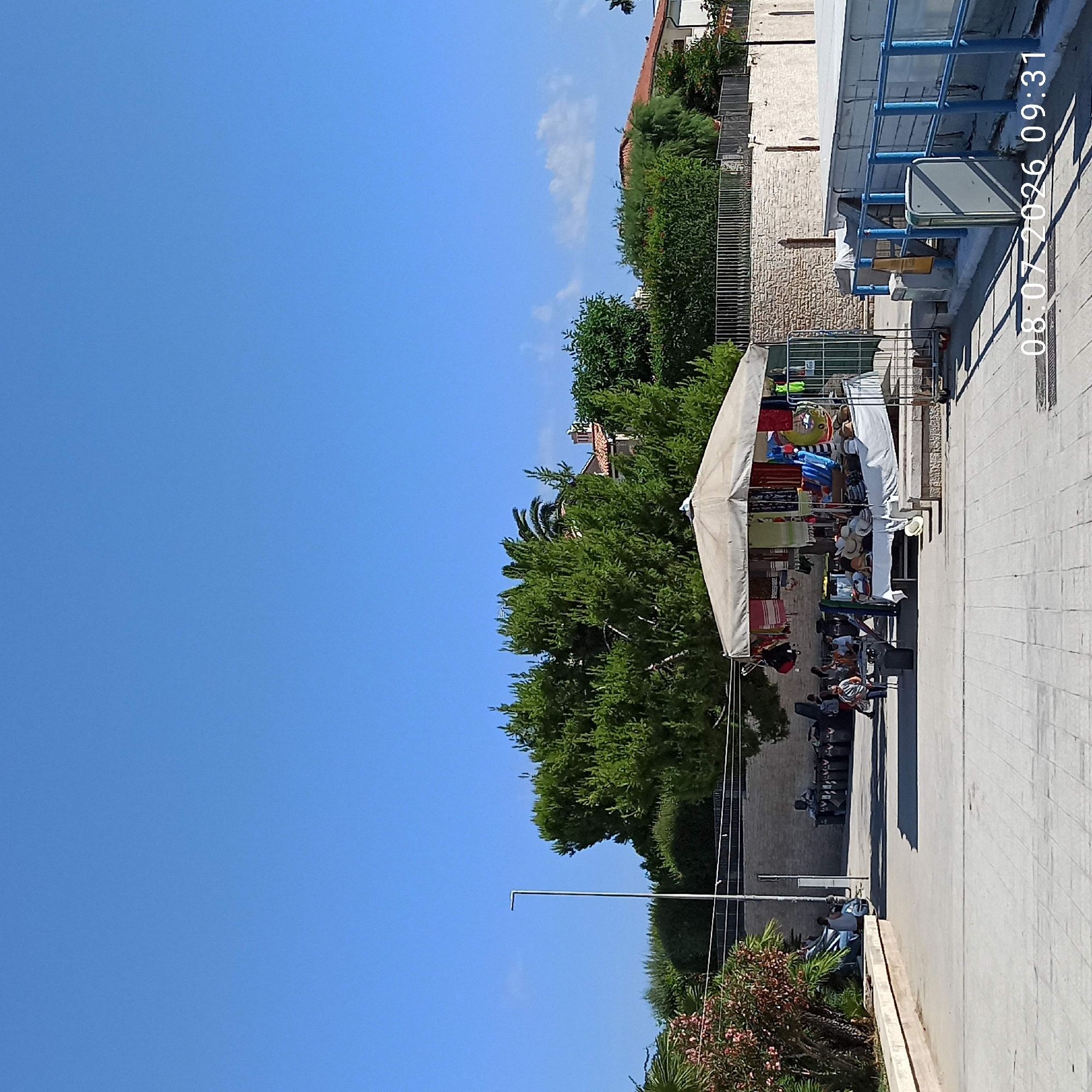 In Trani, Italy, a sunlit outdoor market displays various goods under a large, open-sided canopy. The canopy is draped with colorful textiles and clothing, and a table underneath is covered with hats and other accessories. Several people are browsing the market, and others are visible in the background, near a wall constructed of light-colored stone. Lush green trees and bushes surround the area, with a clear blue sky overhead. A modern lamppost stands to the left of the scene. The ground is paved with light-colored tiles, and shadows are cast by the canopy and other structures. A date and time stamp reads "08.07.2026 09:31".