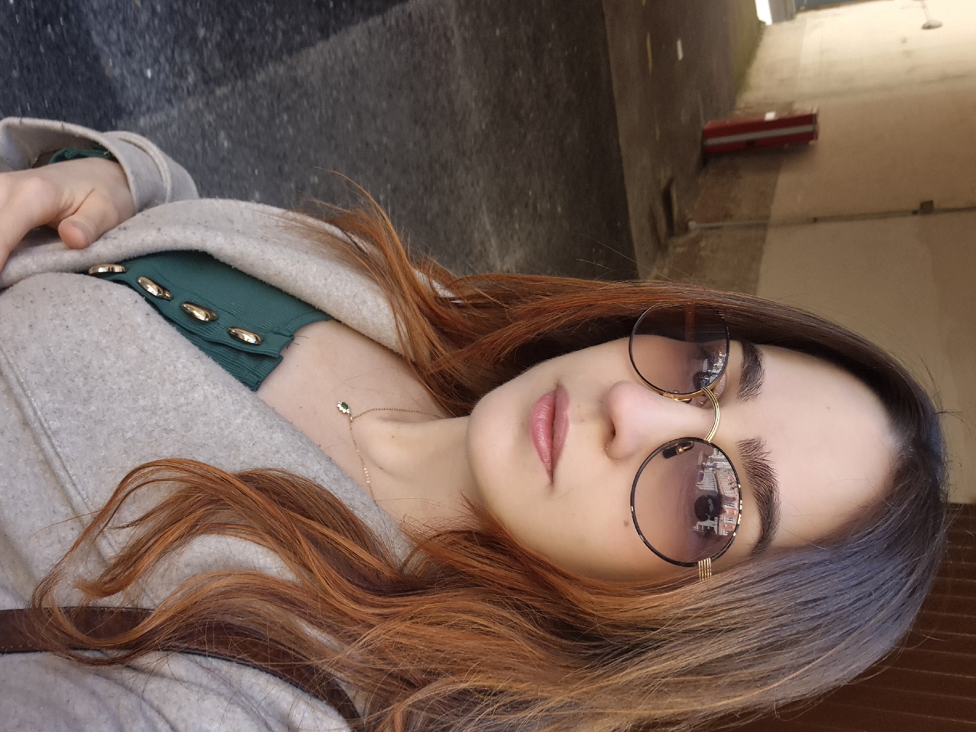 A medium shot shows a person reclining outdoors, likely on a public surface. The person has fair skin, full lips, and is wearing round sunglasses with a gold frame. The lenses reflect the buildings of Strasbourg, France, indicating the location. The person is wearing a green top with gold buttons and a light-colored coat. A gold necklace with a green pendant is visible. The hair is long and ombre, transitioning from brown at the roots to auburn. The background includes a sidewalk and a blurred building facade. A red traffic barrier is situated in the background.