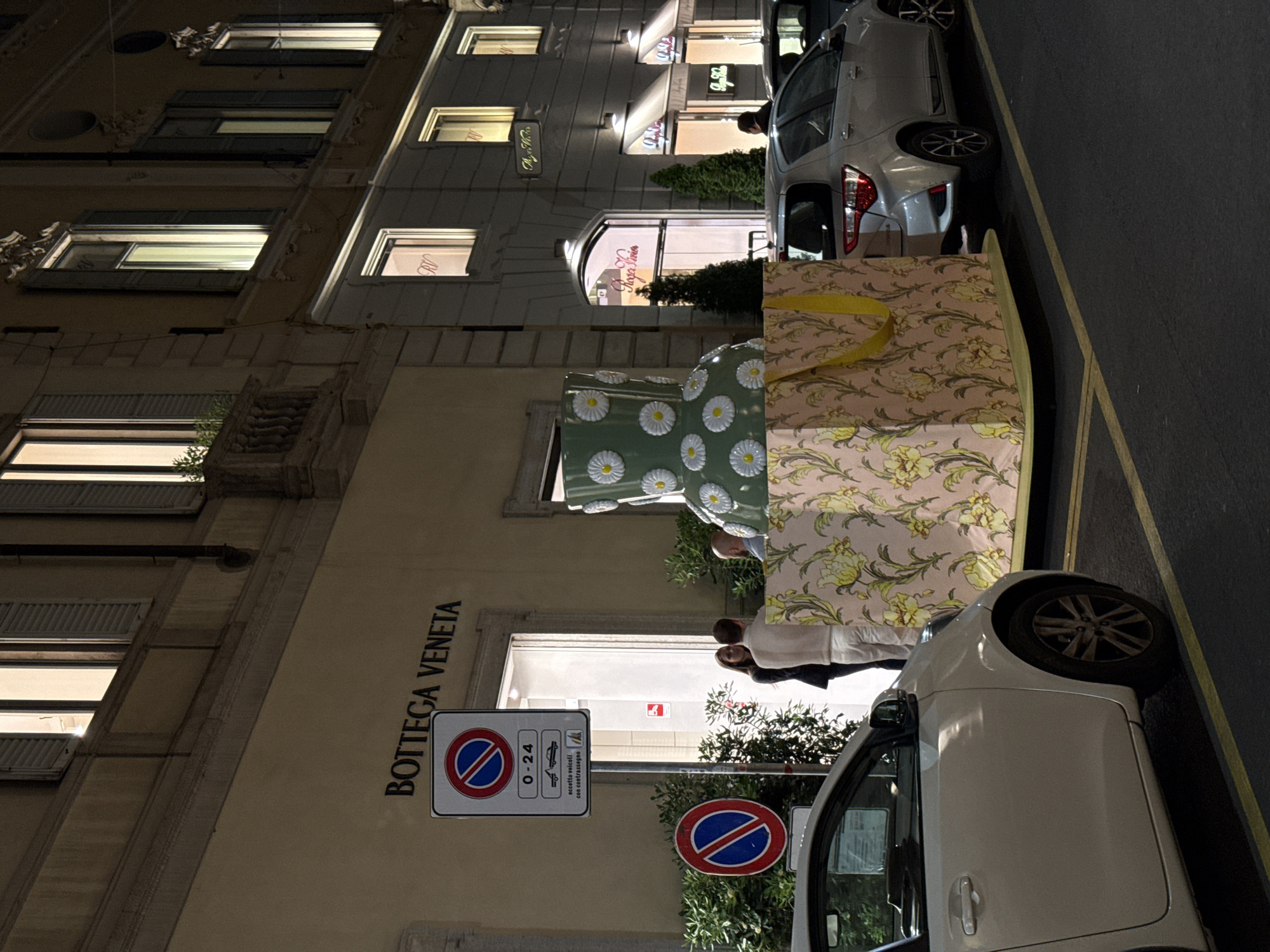 This outdoor nighttime shot depicts a street scene in Milan, Italy, in front of a Bottega Veneta store. Two vehicles, a silver car and a white car, are parked along the curb in front of a street with a yellow curb line. A large, multi-tiered, elaborately decorated display is situated between the parked cars. The building facade features numerous windows, some illuminated, and street signage. People are present near the store entrance. In the background, other businesses with lit signage can be seen. Street lighting illuminates the area.