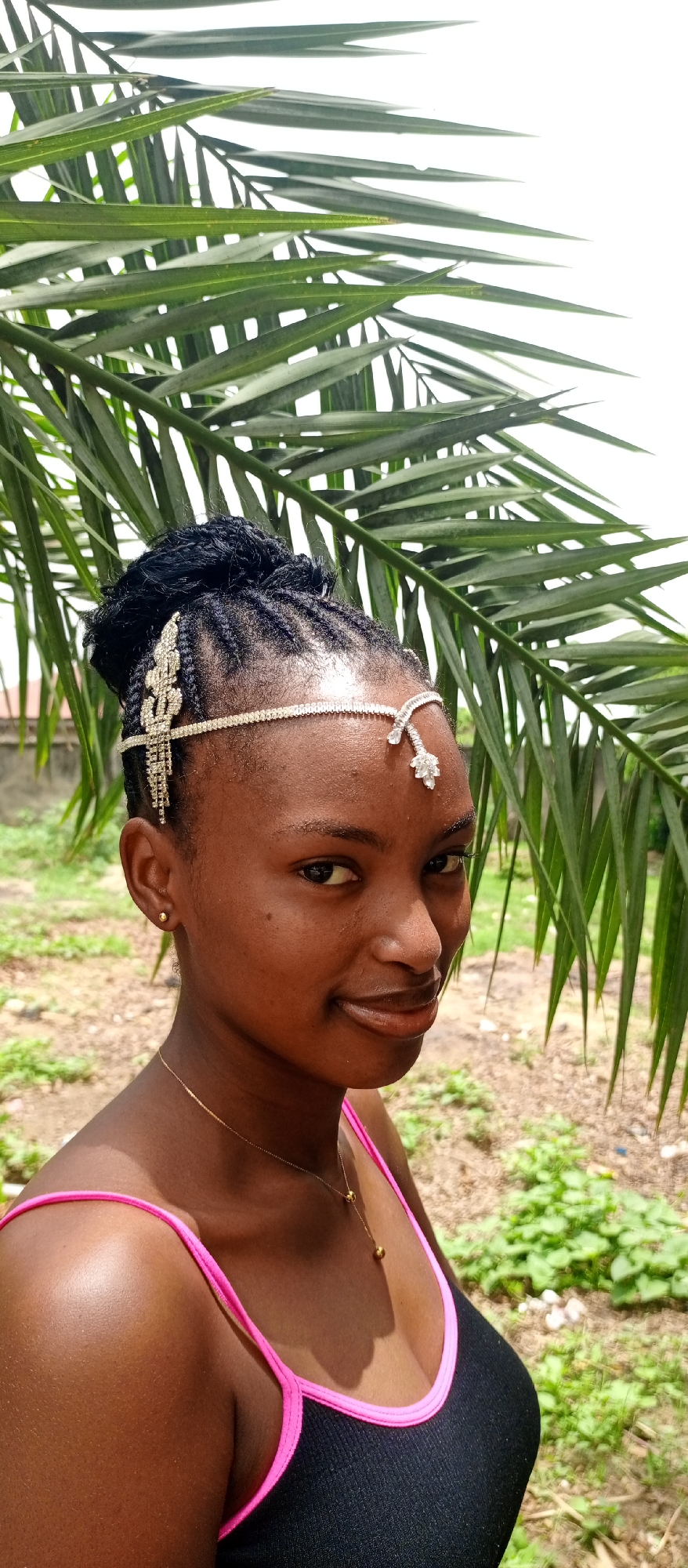 A medium shot captures a young woman with dark skin, likely standing outdoors in Keffi, Nigeria. She is adorned with a jeweled headpiece, her hair styled in braids and pulled up into a bun. Her clothing consists of a black tank top with pink trim. She is wearing a thin, gold necklace. The background shows a palm tree, which suggests the photo was taken in Keffi, Nigeria due to their abundance. The ground is a mix of soil and vegetation.