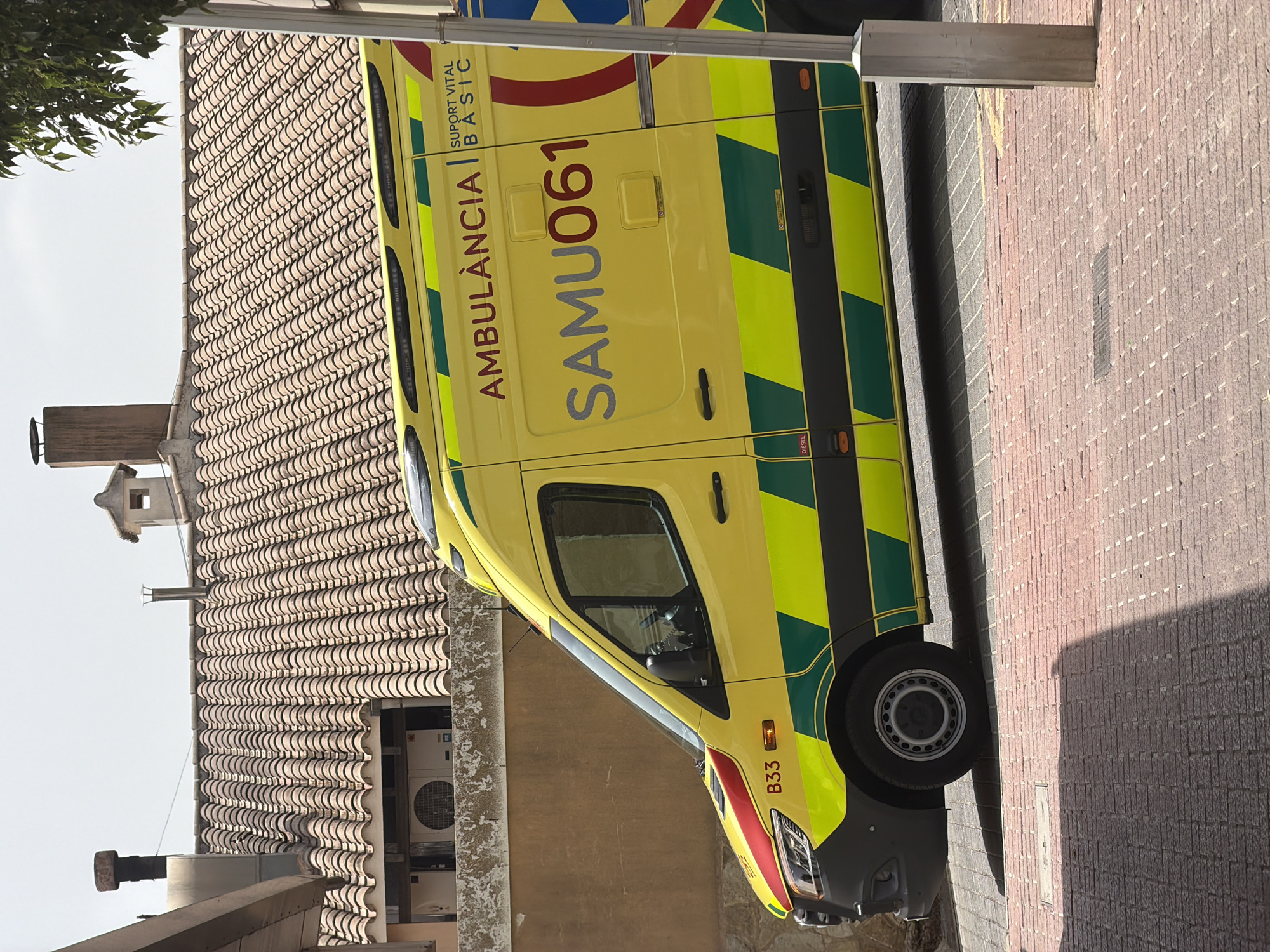 The image shows the side view of a yellow ambulance parked on a street in Palmanova, Spain. The ambulance has "AMBULANCIA" and "SAMU061" written on its side in red and grey letters. The ambulance also features green and yellow reflective stripes. A red and yellow stripe is visible on the front fender, along with the designation "B33". In the background, a building with a tiled roof and a chimney is visible. The street is paved with red bricks. The overall scene is of a parked vehicle in an urban setting.