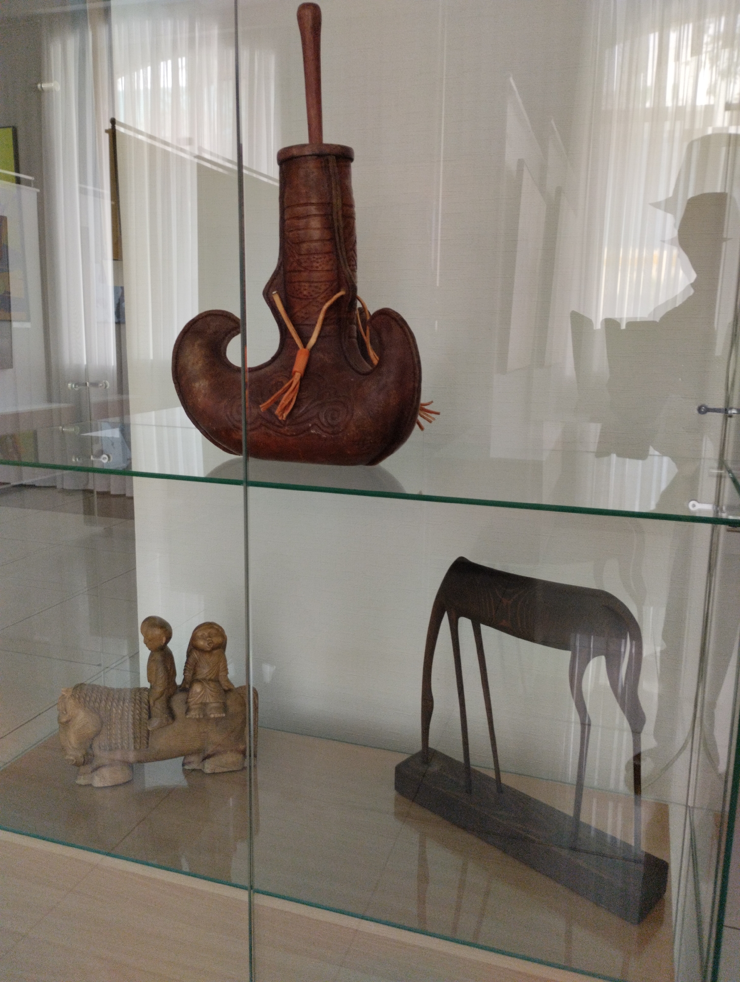 The image shows artifacts displayed in a glass cabinet, likely at the Kostanay Regional Museum of Local Lore in Kostanay, Kazakhstan.
On the top shelf, a large, dark brown, leather vessel with a wooden stopper and decorative orange tassels is displayed. Its shape resembles a stylized boot or horn.
On the bottom shelf, two wooden sculptures are visible. To the left, a light-colored wooden sculpture depicts a horse-like creature with two smaller figures, possibly children, seated on its back. The creature has detailed carvings on its side. To the right, a dark wood sculpture of an antelope or similar animal with long, slender legs stands on a rectangular wooden base.
The background consists of sheer white curtains and a wall, with subtle reflections of a person and architectural elements visible on the glass of the cabinet. The floor appears to be light-colored wood or laminate. The lighting is bright and diffuse, creating some glare on the glass.