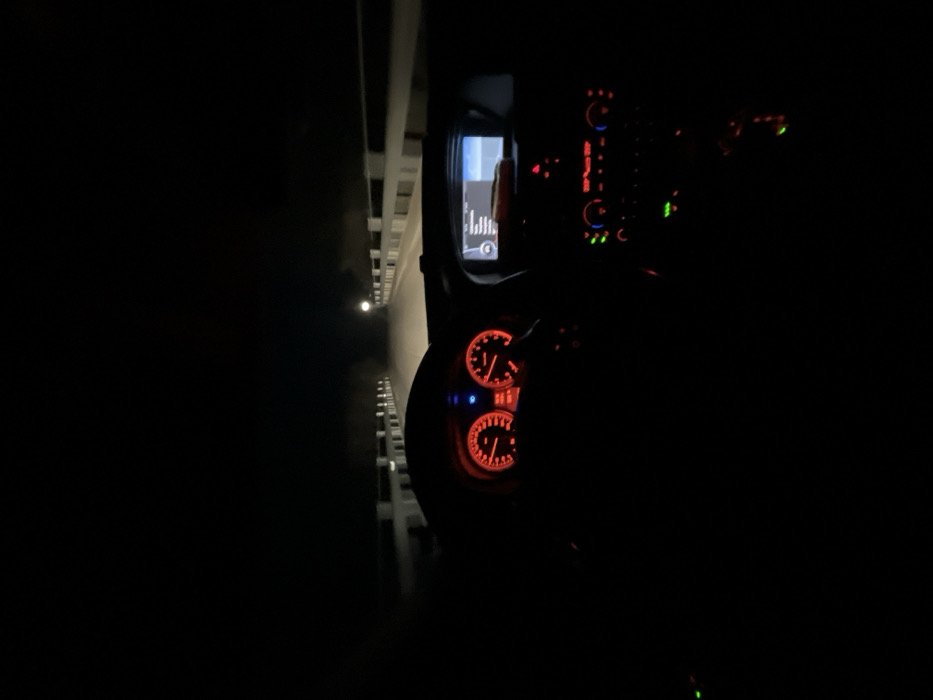 The image depicts the interior of a vehicle at night, observed from the driver's perspective, on a road located in Gradina, Bulgaria. The dashboard instrumentation is illuminated, featuring two primary circular gauges with red-orange backlighting and a blue indicator light. A central infotainment screen, displaying a menu or list, is active to the right of the steering wheel. Below this screen, the climate controls and other buttons are illuminated in green and red. The steering wheel rim is partially visible at the bottom-left. Through the windshield, the vehicle's headlights illuminate a paved road ahead. This road is bordered on both sides by light-colored concrete barrier walls or guardrails that recede into the distance. A single, distinct light source, resembling a street lamp, is visible on the right side of the road in the mid-ground. The distant environment is dark, with some atmospheric haze visible along the illuminated path. No occupants are discernible within the vehicle.