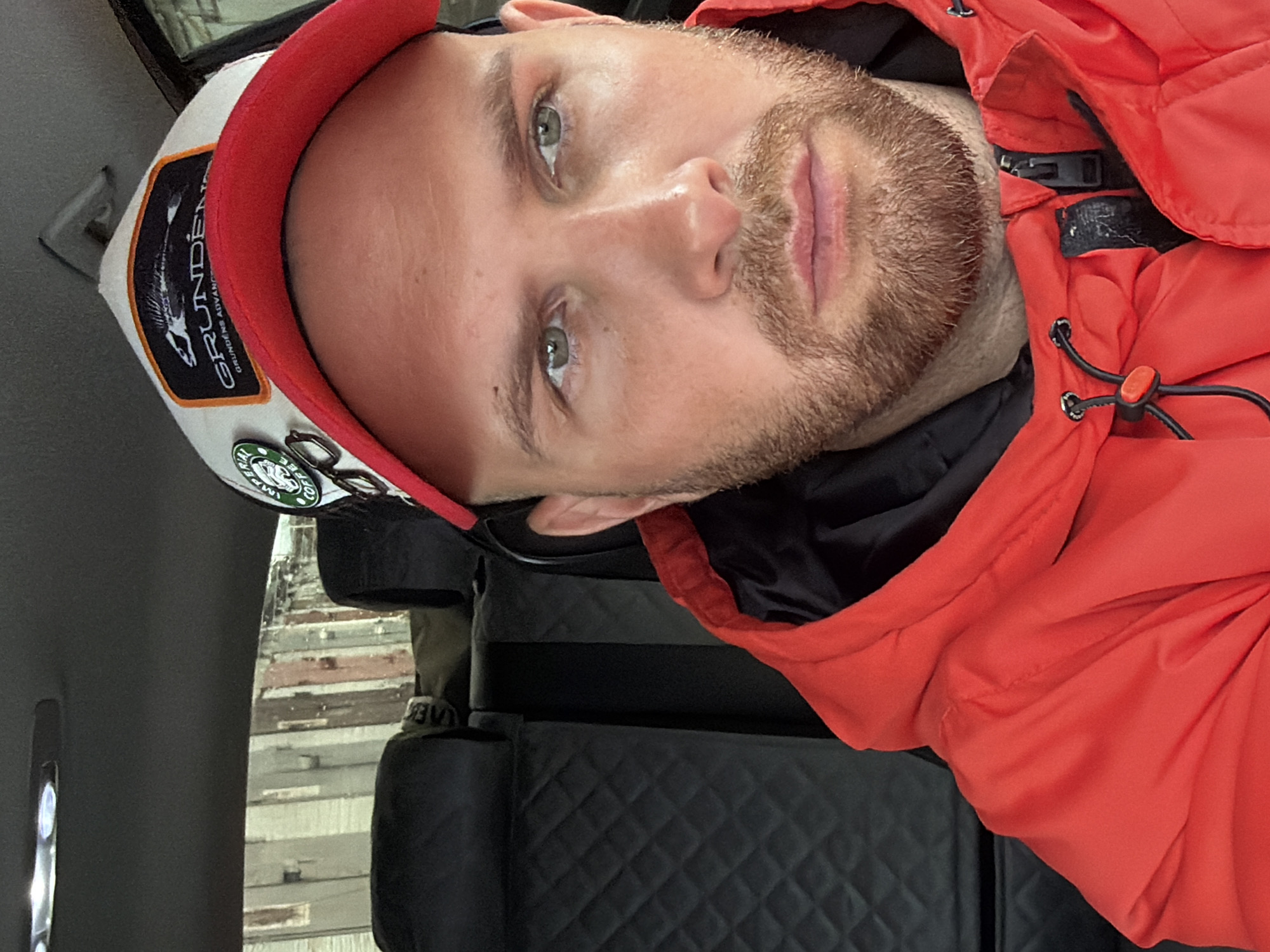 The image features a man with light-colored eyes and a short beard, wearing a white baseball cap with a red brim and a bright red outdoor jacket. He appears to be in an indoor setting, likely a vehicle or cabin, with dark, possibly quilted, seating visible in the foreground. The photograph is taken from an angled perspective, suggesting he might be reclining or the camera is tilted.
On the front of his cap, there is a rectangular black patch with the brand name "GRUNDÉNS" in white text, accompanied by an illustration of a fish skeleton. Below the main brand name, smaller text reads "GRUNDÉNS ALVANYA." Attached to the cap are at least two circular pins, one of which clearly displays the word "COFFEE" in a style consistent with the Starbucks logo. The man's red jacket features black drawstring cords with orange toggles and a black buckle or zipper mechanism near the shoulder.
Through a window in the blurred background, structures resembling wooden planks or siding are faintly visible, suggesting an outdoor environment, possibly an industrial or older building in Chaikovsky, Russia. The man's expression is neutral and calm, and the lighting indicates daylight.