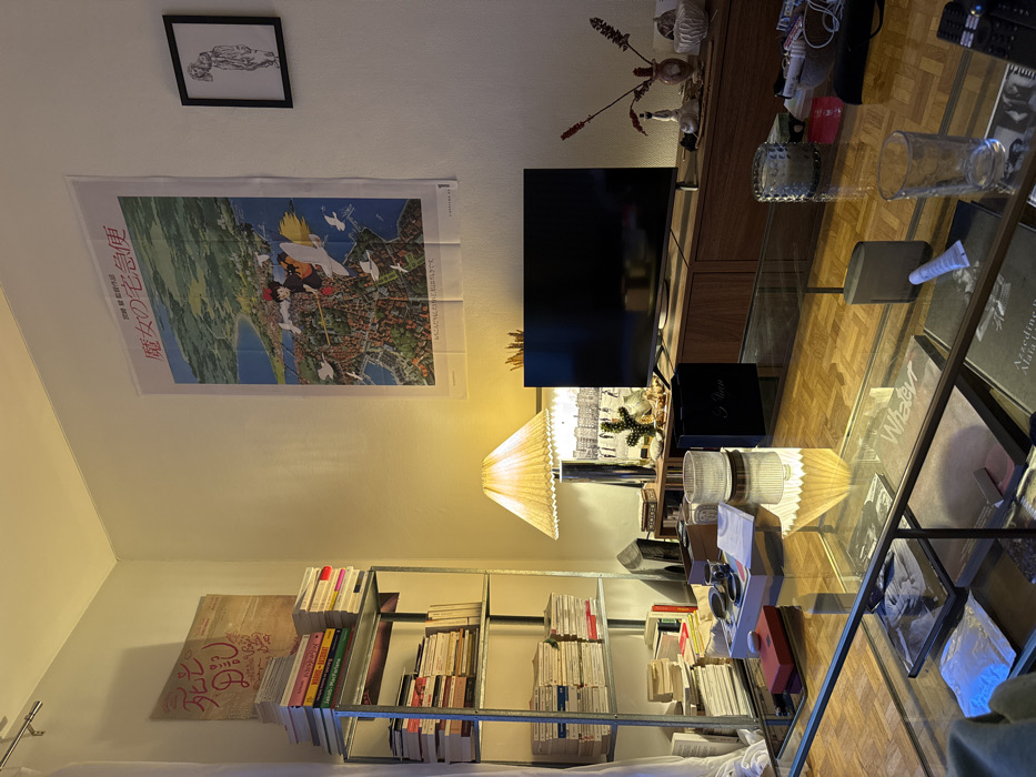 A room features a TV mounted on a cabinet. Above the cabinet, there's wall decor, including a framed picture and a poster. A floor lamp sits near the shelving unit filled with books. A glass-topped coffee table dominates the foreground, displaying various items and reflecting the lamp's light. Hardwood flooring runs throughout the room.