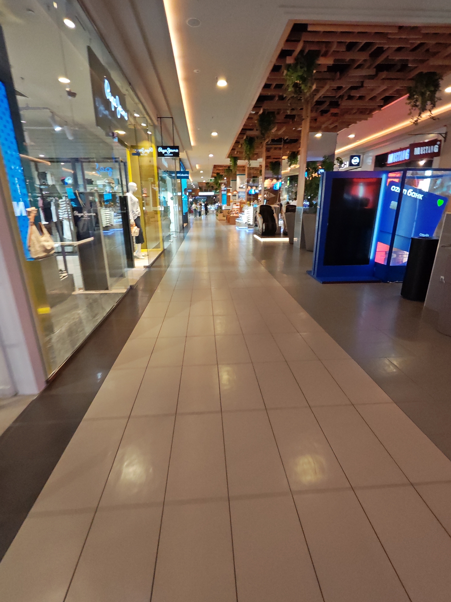 This image depicts the interior of a shopping mall, likely located in Rostov-on-Don, Russia. The perspective is from the floor of a wide, well-lit corridor. The floor is tiled in a light beige color, with dark grout lines forming a grid pattern. To the left, a store window displays clothing on mannequins. The store appears to be a fashion retailer, with visible signage including "Pepe Jeans." The store interior is illuminated, showing racks of clothing and a mannequin dressed in a white shirt and dark skirt.
To the right of the corridor, there are large, illuminated digital screens. One screen is predominantly blue and displays text in Cyrillic script, possibly advertising a bank or financial service, with the visible text "oz n банк." Further down the corridor, another screen is dark. Above the digital screens, the ceiling is adorned with a decorative wooden lattice structure, from which artificial greenery hangs. Above the left-side store, the ceiling is white with recessed lighting.
In the distance, several people are walking through the mall. The overall atmosphere is clean, modern, and commercial. The lighting is bright, creating reflections on the polished floor.