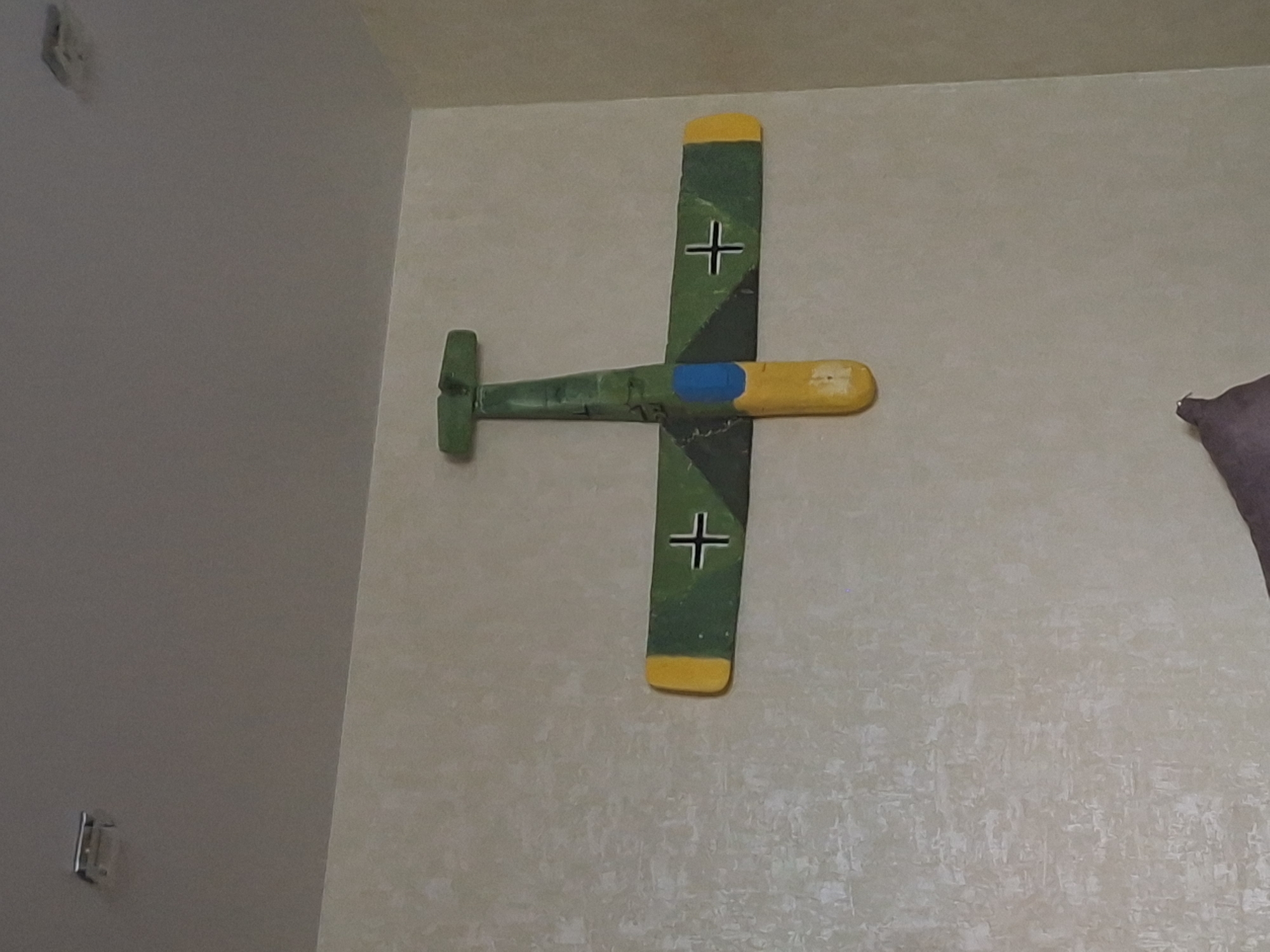 A wooden model of a German World War II aircraft is mounted on a light-colored wall. The model is painted with green camouflage, yellow highlights on the fuselage and wingtips, and black crosses. The model is horizontally oriented with the tail on the left side of the image. The wall behind the model is textured. The location is the city of Serpukhov, Russia.