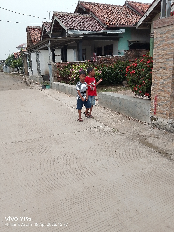 The image shows two young boys standing on a paved road in Anjatan, Indonesia, located at latitude -6.412 and longitude 107.962. The road is lined with houses, some of which have red tile roofs. There are bushes with red flowers and a brick wall in the background. The boy on the left is wearing a black and white striped shirt and blue shorts, while the boy on the right is wearing a red shirt and blue shorts. The boys are looking at something in the distance. The date and time are stamped on the bottom right of the image: "18 Apr 2025, 13.37."