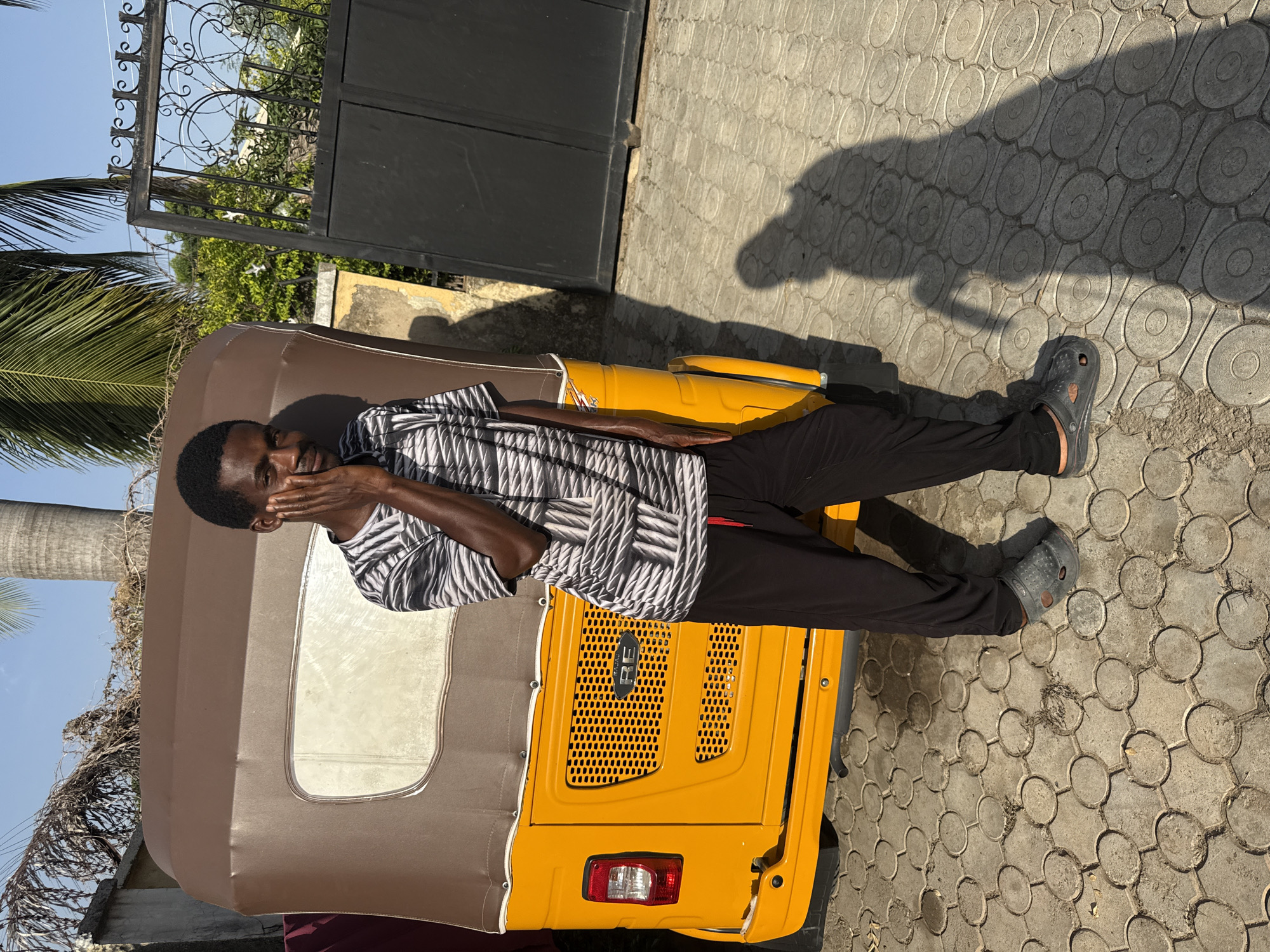 The image displays a young man standing next to a yellow and brown auto-rickshaw on a brightly lit day in Abuja, Nigeria. The man, who appears to be of African descent, wears a black and white patterned short-sleeved shirt, black trousers, and dark grey clogs. He stands with his right hand partially covering his mouth and nose, looking directly at the camera, conveying a sense of shyness or a suppressed smile.
The auto-rickshaw, a common mode of transport, features a vibrant yellow body and a brown, soft-top canopy. On its rear, a black metallic grille is visible, centered with an oval-shaped logo that appears to read "KERE". A single red taillight is also visible on the back. The vehicle is parked on a paved surface composed of light grey interlocking stones, arranged in a circular pattern.
In the background, a tall, dark metal gate with decorative wrought-iron elements is seen, partially framed by green foliage and what appears to be a palm tree on the left side of the frame. The strong, high sunlight casts distinct, elongated shadows on the ground, indicating it is likely midday or early afternoon with clear weather conditions. The overall scene captures a casual moment in an urban or semi-urban setting.