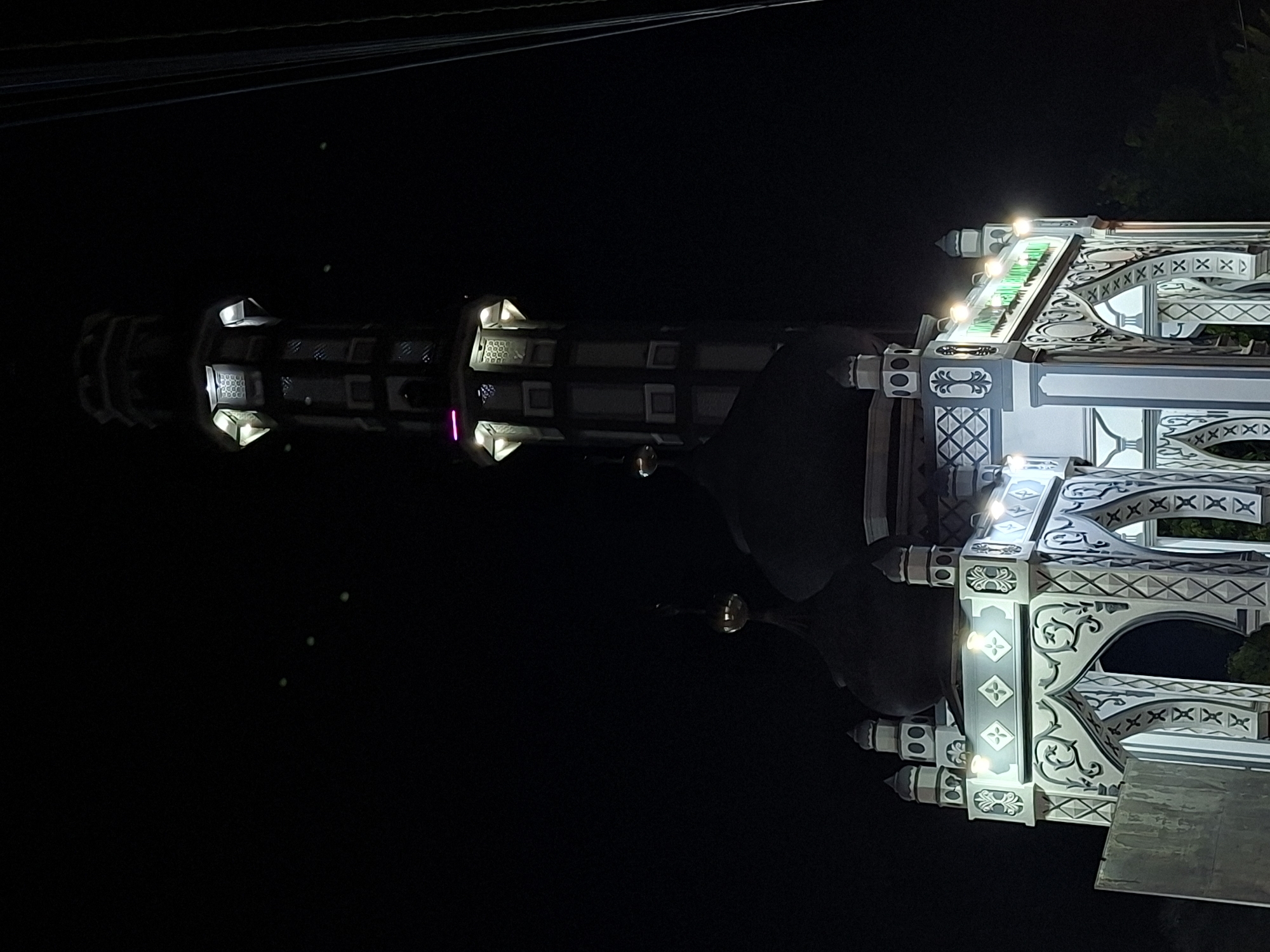 A low-angle, long-exposure photograph taken at night depicts a structure, likely a mosque, located in Keudekruenggeukueh, Indonesia. The building features detailed architectural ornamentation in white and gray, illuminated by multiple light sources. A minaret is visible extending vertically into the dark night sky. Overhead power lines are visible against the top of the frame.