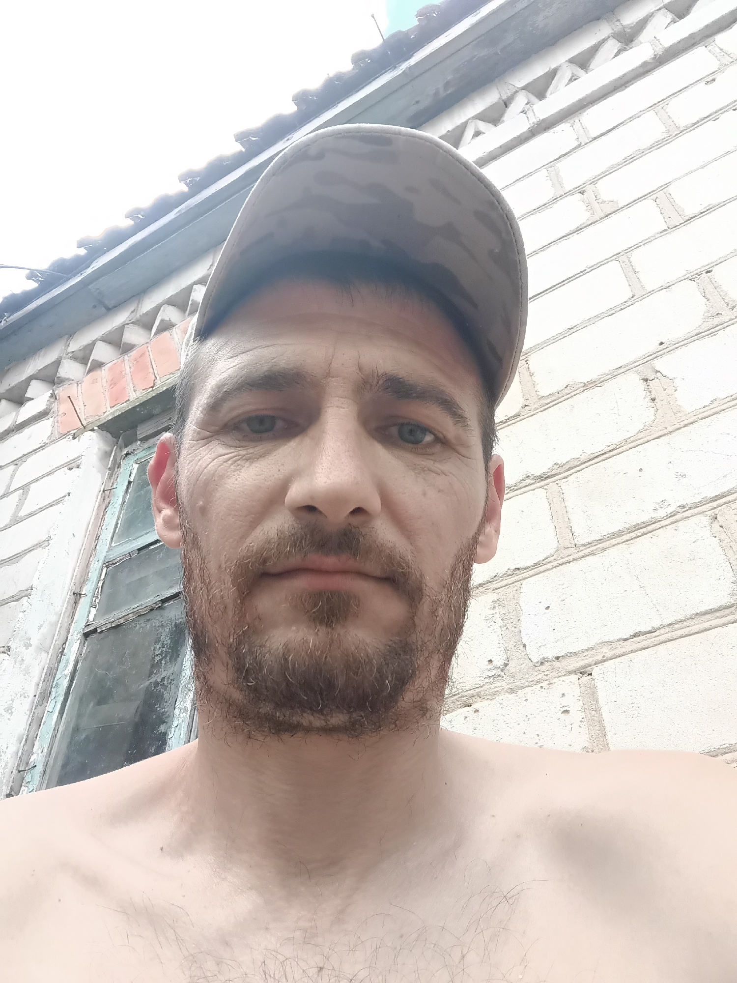 The media file is a close-up, low-angle selfie of a shirtless man wearing a camouflage baseball cap. He has short, brown hair and a light beard. His eyes are blue and he has a neutral expression. The background shows a white brick wall with some signs of wear and tear, and a portion of an old, weathered window with peeling blue paint. Above the wall, a section of a roof is visible, with some sunlight filtering through. The overall setting appears to be outdoors, possibly in a rural or older urban area. The lighting suggests it is daytime, with bright, overcast conditions. There are no visible text elements in the image. The location context provided is Saksahan, Ukraine.