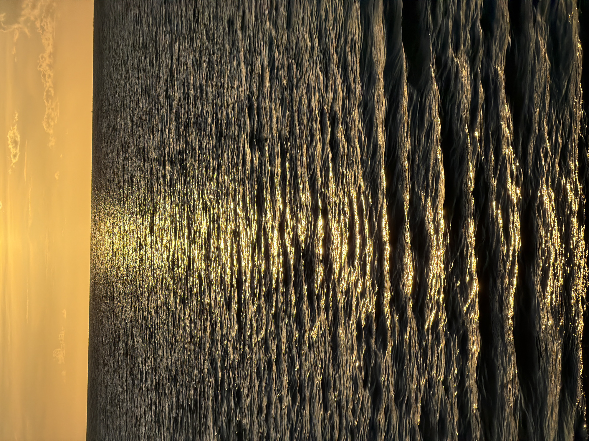 This is a photograph of the sea at sunset. The sky is a vibrant orange, with a few wispy clouds. The sun's reflection creates a golden path across the rippling water. The scene is devoid of any people, structures, or landmasses, suggesting an open body of water. The image is taken from a low angle, emphasizing the expanse of the sea. There is no information available about the specific location in Baan Plai Laem, Thailand, other than that it is a coastal area.
