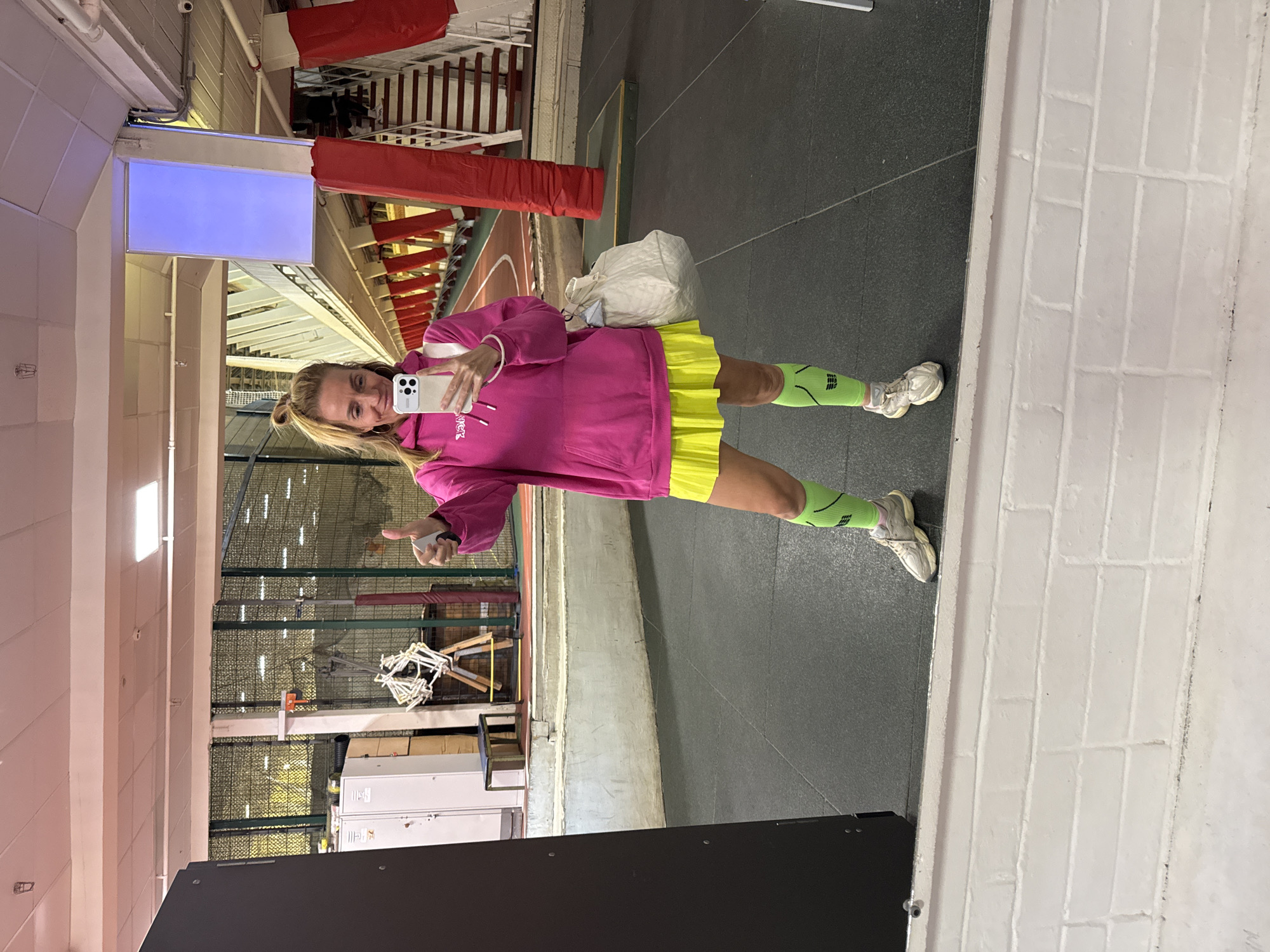 A woman stands in an indoor sports facility, reflected in a mirror. She is wearing a pink hoodie, a yellow skirt, green socks, and white sneakers. She is holding a white mobile phone and posing with a peace sign. She also has a white bag on her shoulder. The background reveals a gymnasium with sports equipment and spectator seating. The city of Saint Petersburg, Russia is not discernible from the image.