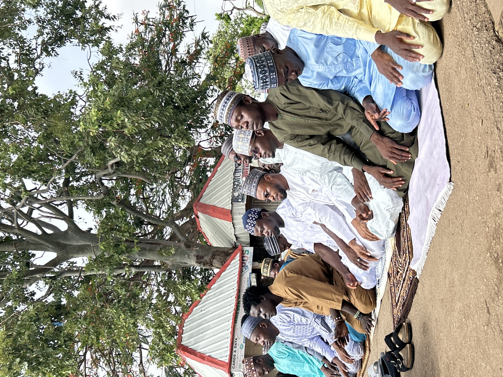A group of approximately a dozen men wearing a variety of traditional Muslim attire are praying outdoors. They are kneeling in rows on a patterned rug placed on the ground. The men are predominantly young, with various skin tones and wearing different colored clothes, including white, green, blue, and brown. Many wear small embroidered hats. A large tree with dense foliage is behind the praying group. Behind them is a small building with a red and white striped awning where signs for "CARDS & DATA" are displayed. This scene is likely in Jos, Nigeria.