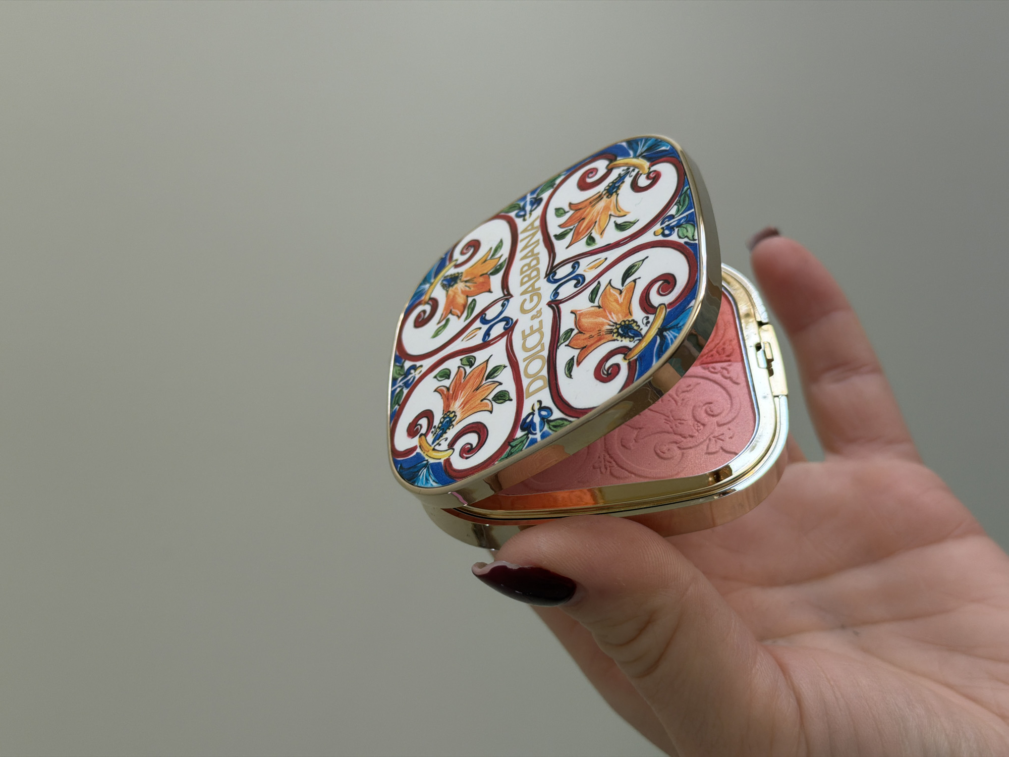 A hand with dark manicured nails holds a Dolce & Gabbana blush compact against a neutral grey background. The compact is open, revealing a rose-colored blush with a subtle floral pattern pressed into the surface. The lid of the compact features a decorative, colorful tile-inspired design with floral motifs and the Dolce & Gabbana logo. The compact has a gold-toned frame. The location is not discernible from the image and cannot be confirmed as Dreiliņi, Latvia.