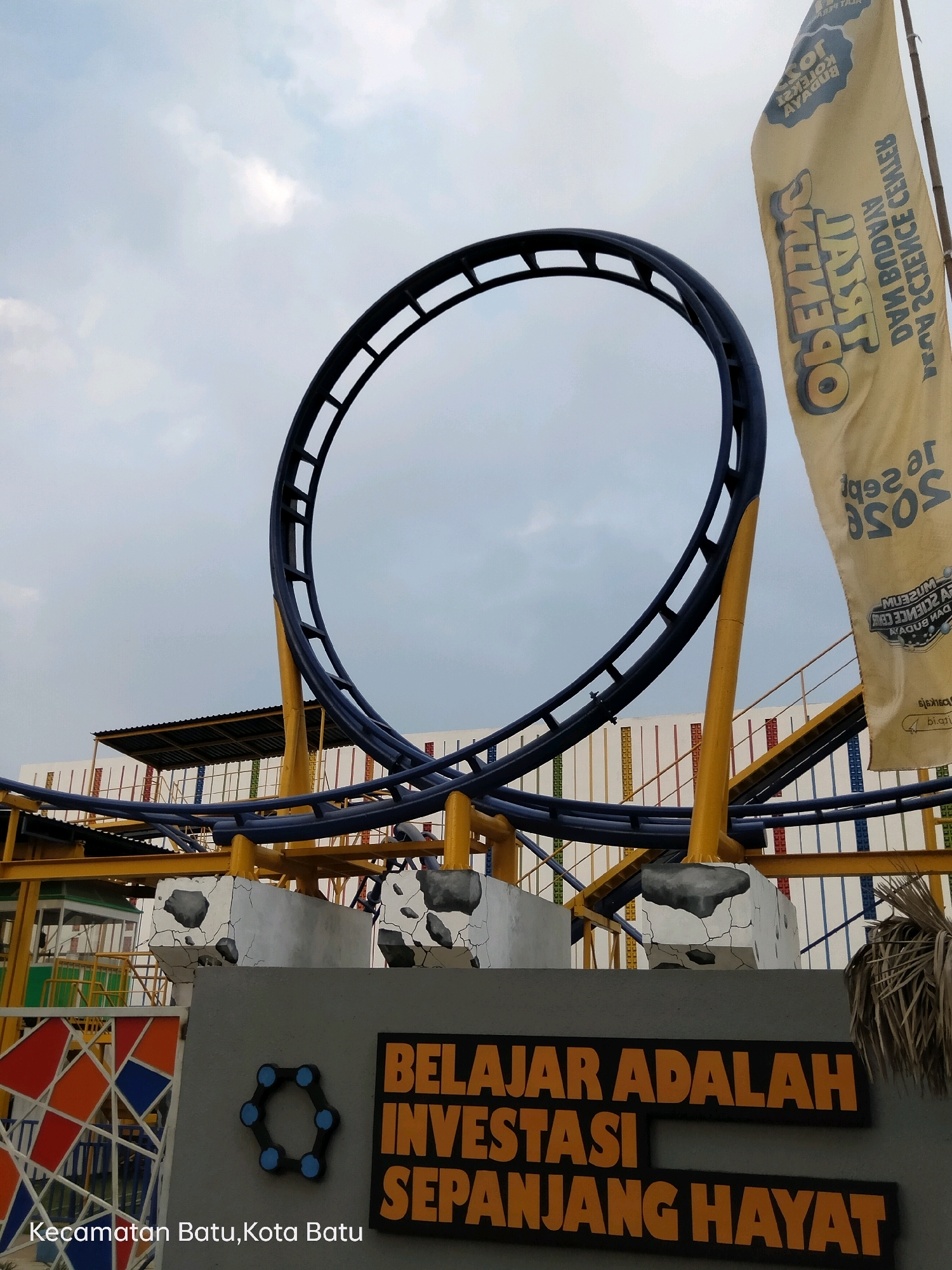 The image shows an amusement park with a blue roller coaster track forming a loop against a cloudy sky. A yellow flag with Indonesian text and graphics is prominently displayed on the right side. In the foreground, a grey wall features the text "BELAJAR ADALAH INVESTASI SEPANJANG HAYAT" in orange letters, which translates to "Learning is a lifelong investment." Below this, the location is identified as "Kecamatan Batu, Kota Batu," indicating it is in the Batu District, Batu City, Indonesia. Various decorative elements and structural supports of the roller coaster are visible, along with a colorful geometric fence on the left. The overall scene depicts an outdoor recreational facility.