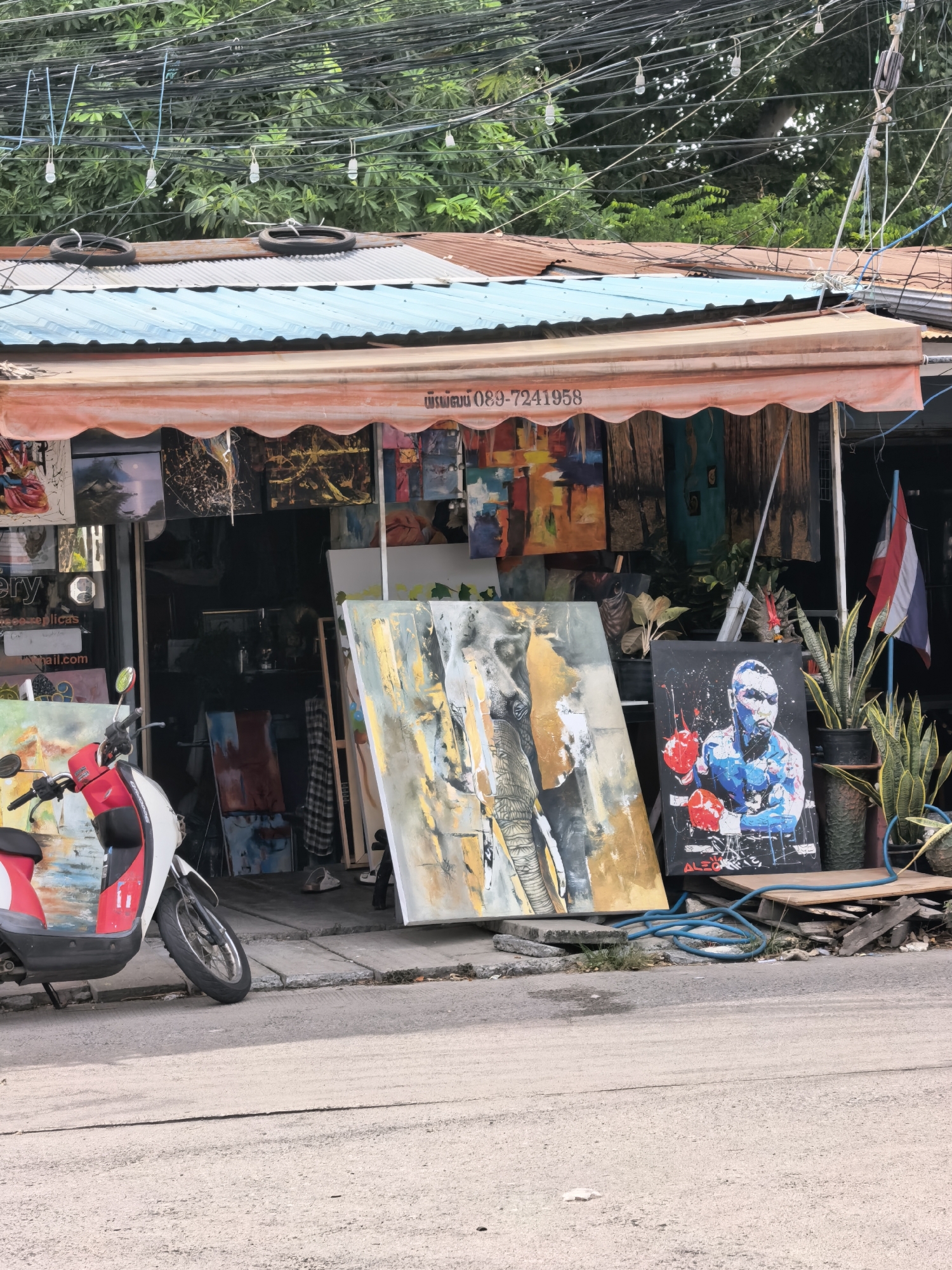 This image depicts an outdoor art gallery or shop in the Ko Samui District of Thailand. The storefront features an orange awning with a scalloped edge, under which various paintings are displayed. Some paintings hang on the wall of the shop, while others are propped up outside on the ground. Prominently featured are a large abstract painting with elephant imagery and another painting of a boxer. A red and white scooter is parked to the left of the entrance. The shop is situated along a street with visible power lines overhead and lush green foliage in the background. A Thai flag is visible on a pole to the right of the shop. The scene suggests a commercial establishment in a tropical urban environment.
