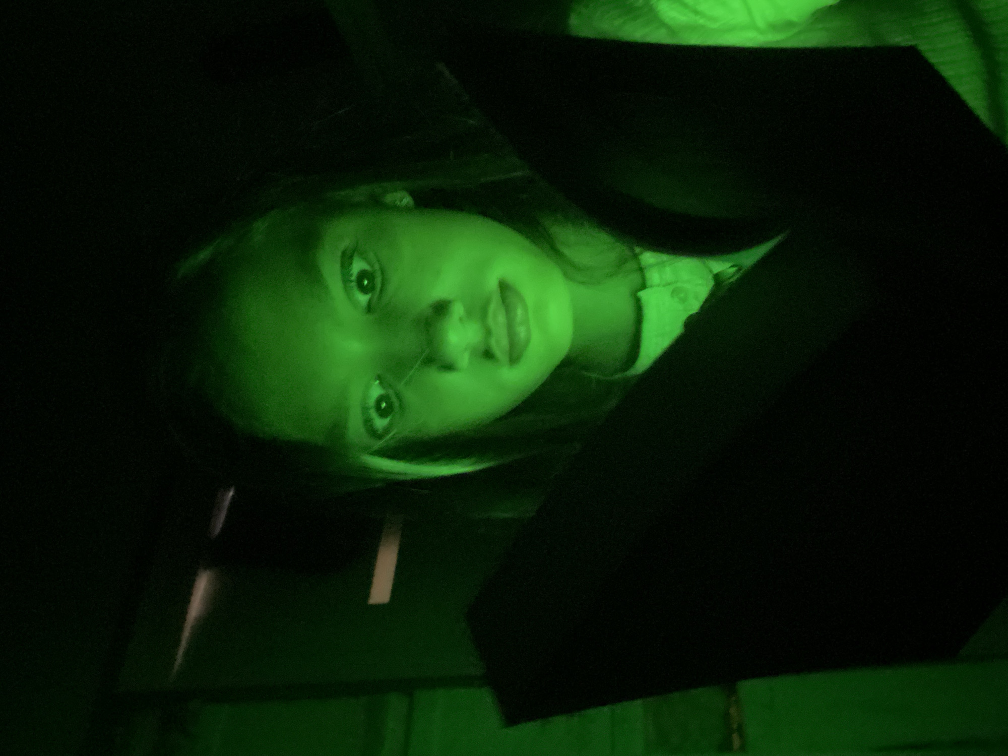 A young girl with wide, startled eyes stares directly at the camera in a dimly lit environment, illuminated by a harsh green light. She appears to be in an enclosed space, possibly a car, given the dark shapes that suggest the interior of a vehicle. The girl's face is the primary focus, with her hair falling around her face. Dark, abstract shapes are prominent in the foreground, obscuring parts of the image and contributing to a slightly unsettling atmosphere. The overall impression is that of a candid, perhaps spontaneous, photograph captured in a moment of surprise or fear, with the green light casting an unnatural glow on the scene. The location is not specified in the image or the provided crops, therefore the city of Novosibirsk, Russia cannot be determined.