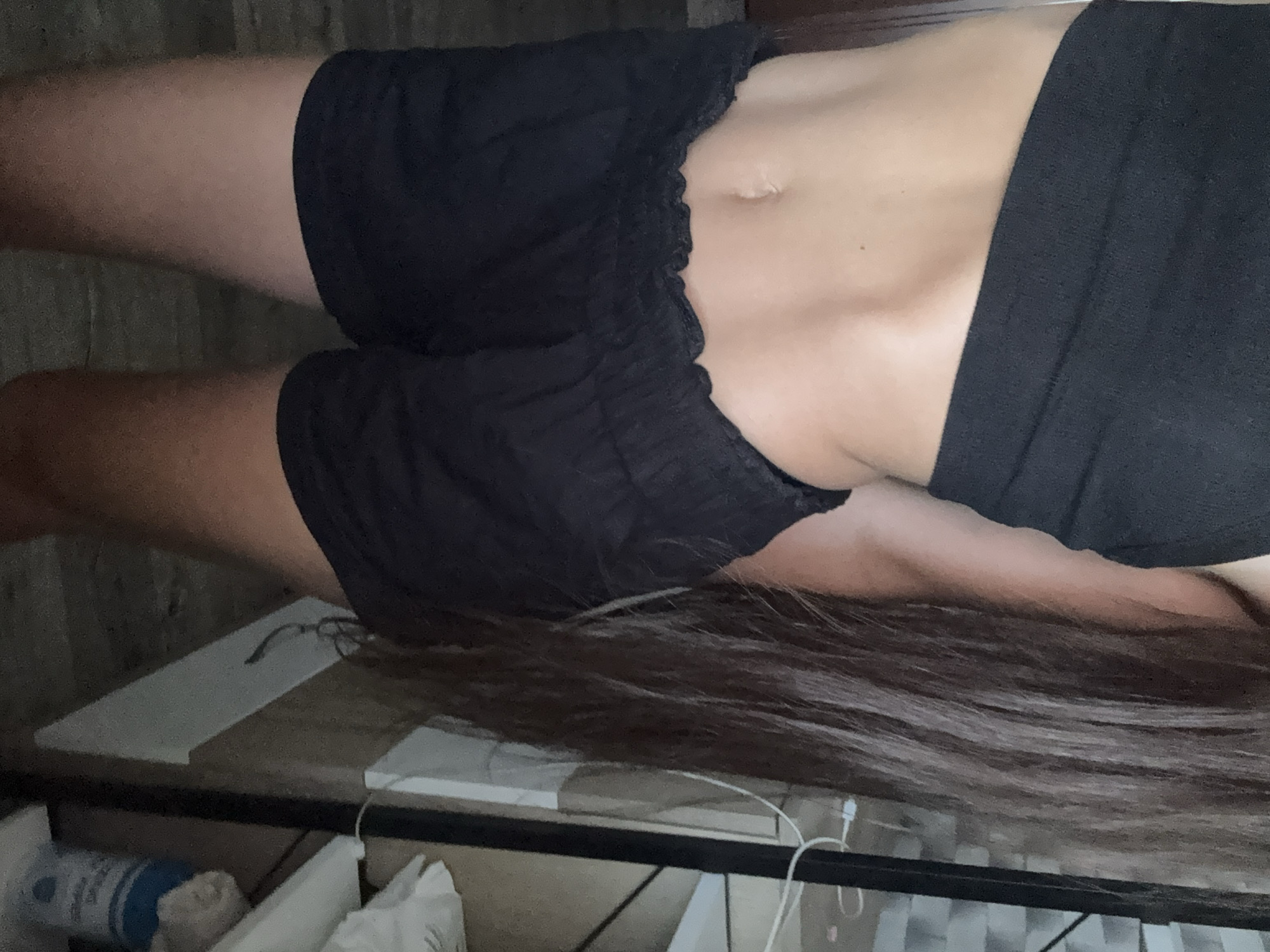 The image shows a close-up of a person's torso and legs, wearing a black crop top and black shorts. The person has long, dark brown hair that extends down their right side. Their midsection is visible, showing toned abdominal muscles. The background appears to be an indoor setting, possibly a room, with a shelf and some items on it, including a blue container. There are no visible indications of the specific location within Basarabeasca, Moldova.