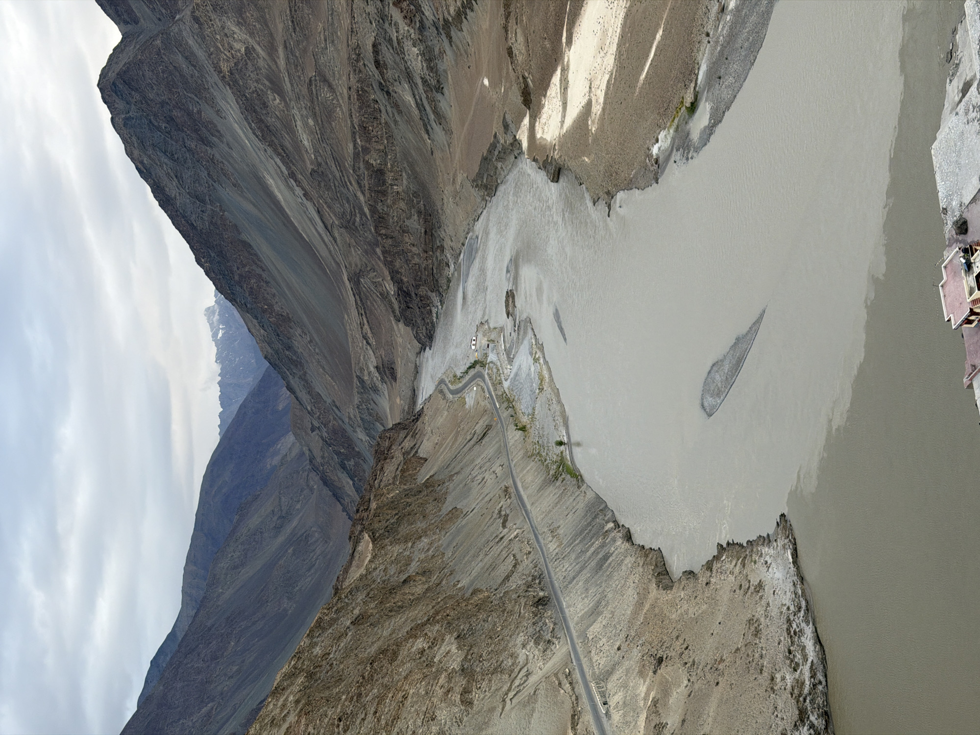 A high-angle, vertical shot captures a wide, shallow river flowing through a rugged, mountainous valley. The river's water is a milky, light brown, with darker, more turbulent currents visible where two bodies of water appear to merge. On the left bank, a winding paved road hugs the steep, rocky slope, which is covered in loose scree and sparse vegetation. A small, white building with a red roof is visible at the bottom right, situated on a paved area near the riverbank. The valley walls are steep and barren, showing layers of sedimentary rock formations. In the distance, the valley narrows, revealing more rugged peaks under a cloudy, overcast sky. The scene depicts a remote, natural landscape with minimal human presence, suggesting a location like the confluence of the Indus and Zanskar rivers near Leh, Ladakh, India, an area known for its dramatic mountain scenery and glacial rivers.