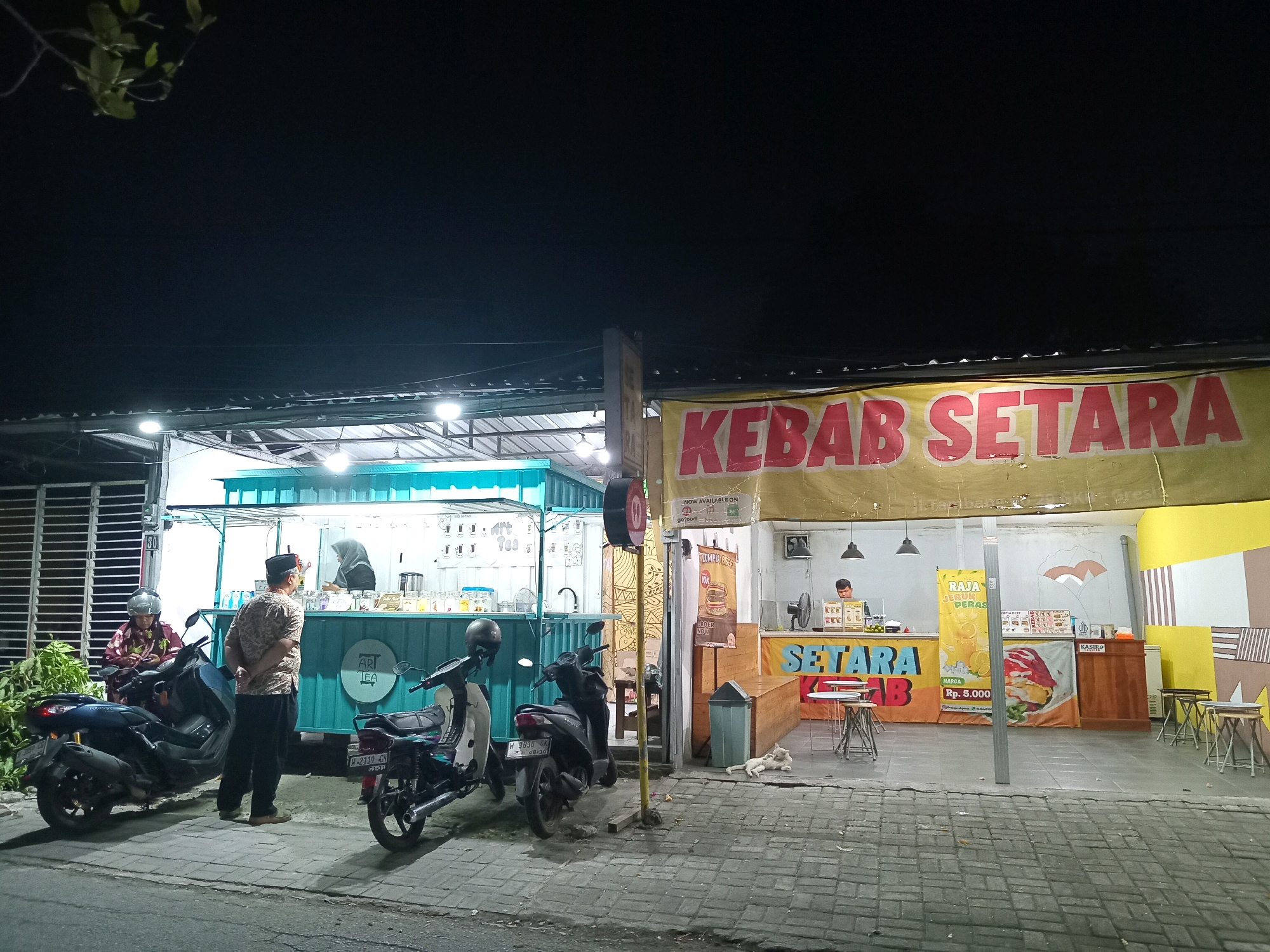 The image depicts an outdoor scene at night in Randuagung, Indonesia, featuring a food stall and a cafe. The main focus is on a food stall with a large yellow banner displaying "KEBAB SETARA" in red lettering. To the left of the kebab stall, there is a teal-colored container-like structure that appears to be a tea shop named "Ari Tea".
In front of the tea shop, two individuals are visible. One person is seated on a dark-colored motorcycle, wearing a patterned shirt and a peci hat, looking at their phone. Another person, in a patterned shirt and dark pants, stands with their hands behind their back, facing the tea shop. Two motorcycles are parked on the tiled sidewalk in front of the shops.
In front of the kebab stall, a man is working behind the counter. A poster advertising "Raja Jus Jeruk Peras" (King Squeezed Orange Juice) for Rp. 5,000 is displayed. The stall has seating arrangements with small tables and stools. A white cat is lying on the pavement in front of the stall. The environment appears to be a street or a small market area. The lighting is artificial, coming from the shop signs and overhead lights.