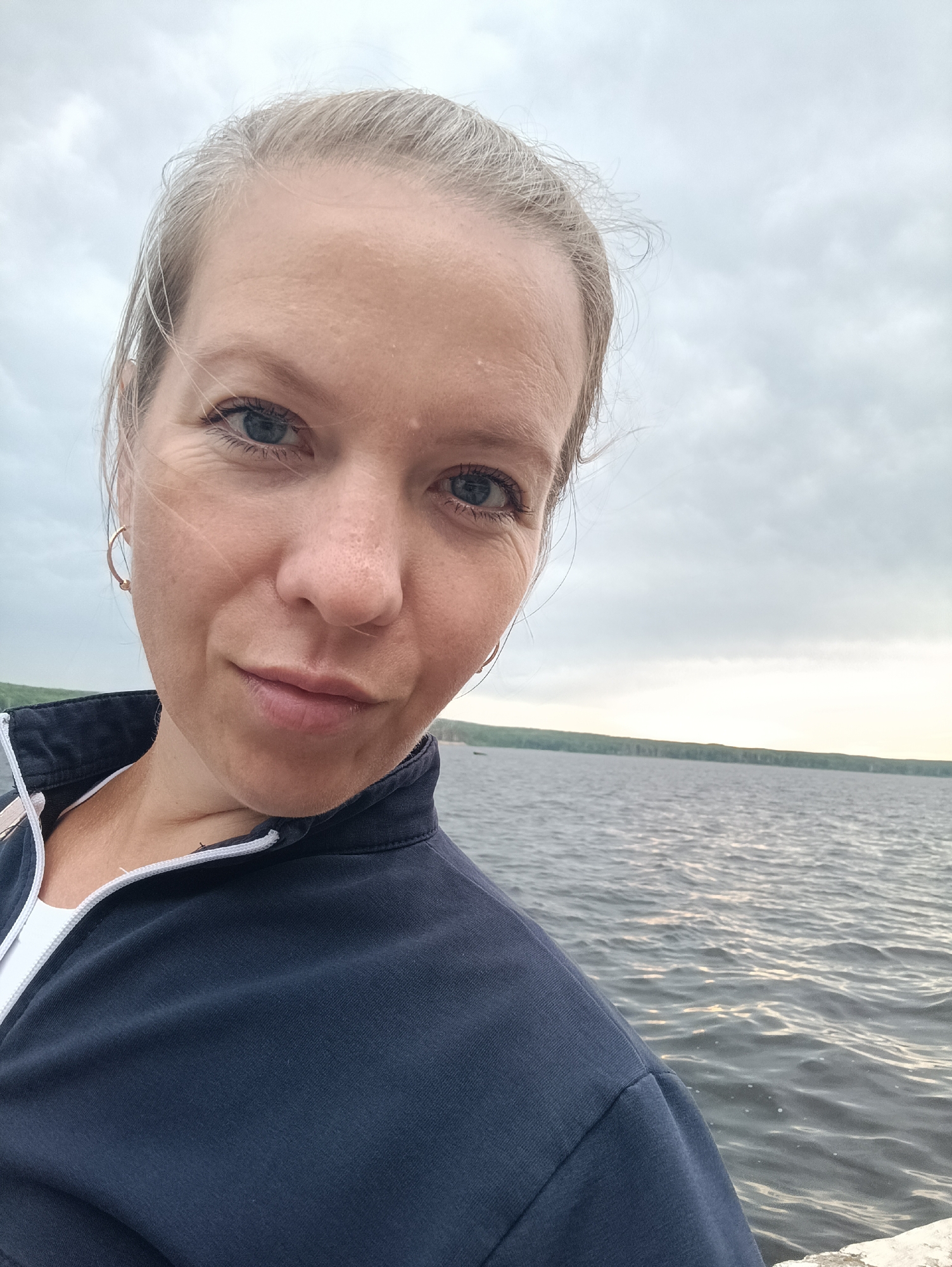 A selfie of a fair-skinned woman with blonde hair pulled back. She is wearing a dark blue zip-up jacket over a white shirt. She is looking at the camera with a neutral expression. Behind her, there is a body of water, likely the Kama River, with a treeline in the distance. The sky is overcast. The image appears to have been taken outdoors in Chaikovsky, Russia, likely near the Kama River embankment.