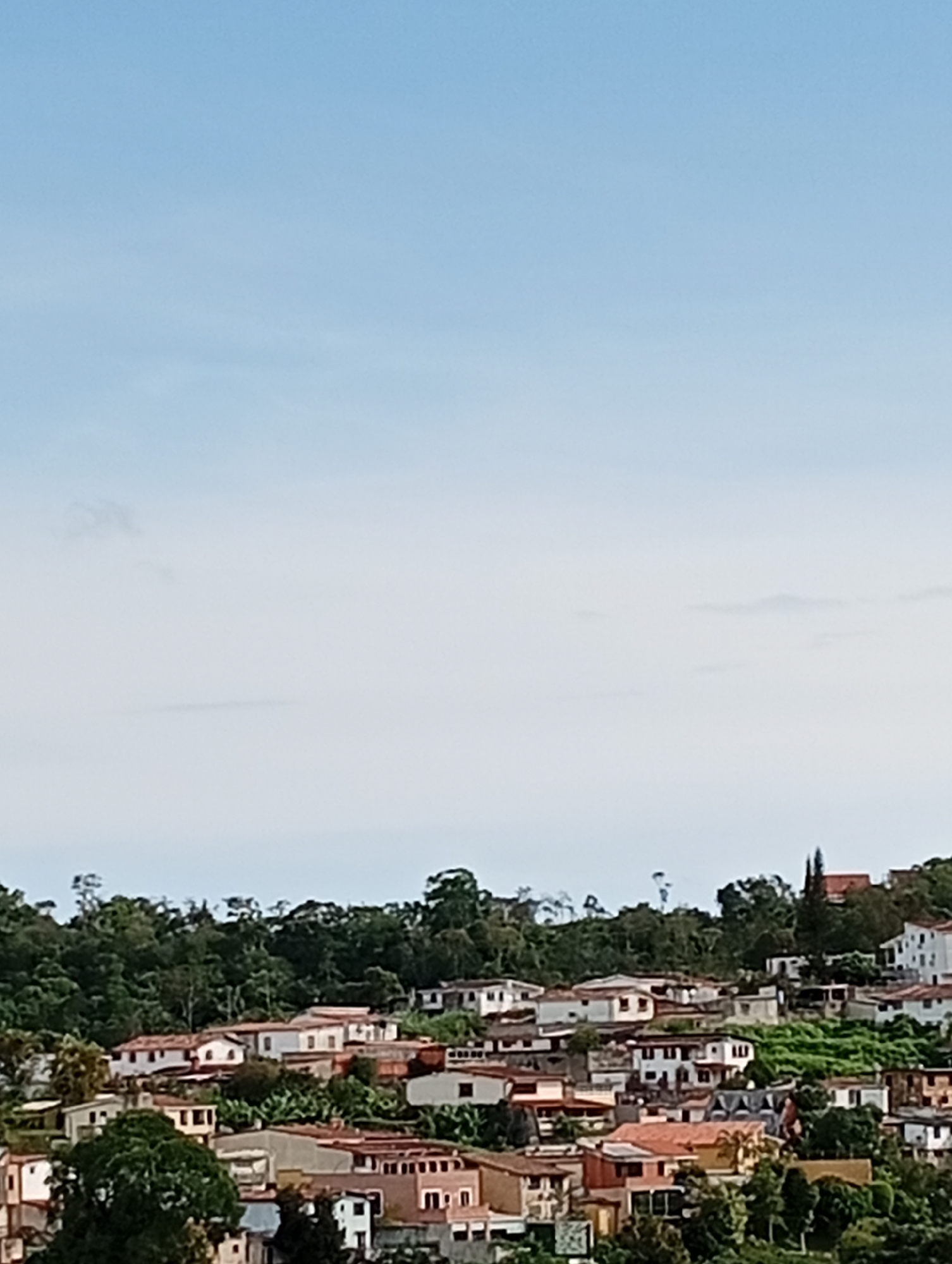 A medium shot captures a residential hillside in San Antonio de Los Altos, Venezuela, under a bright, partly cloudy sky. The scene is dominated by a dense cluster of houses with red-tiled roofs, nestled among lush green trees and vegetation. The houses vary in size and architectural style, with some appearing older and others more modern, their white and earth-toned walls peeking through the foliage. The terrain is uneven, with houses built at different elevations, creating a picturesque yet dense urban landscape. No people are visible in the image, and the focus is entirely on the cityscape and its natural surroundings.