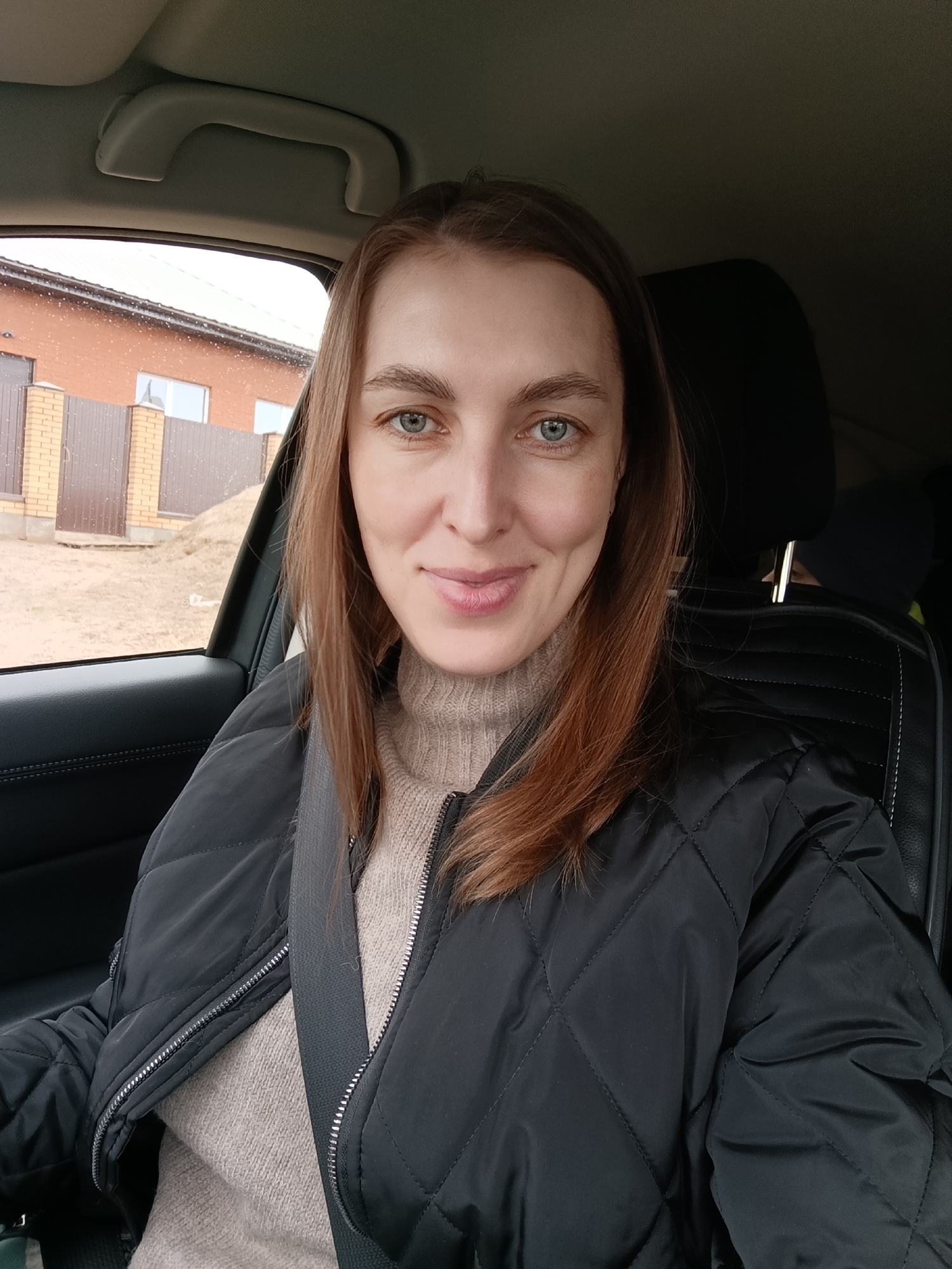 A woman is sitting in a car, wearing a black quilted jacket over a light brown turtleneck sweater and a seatbelt. She has fair skin, light blue eyes, and reddish-brown hair. Outside the car window, there is a two-story brick building with white-framed windows and a brown fence. There's also some light brown soil or grass visible, with some possible snow or light rain visible on the window. The scene is in an unspecified area of the city of Chaikovsky, Russia.