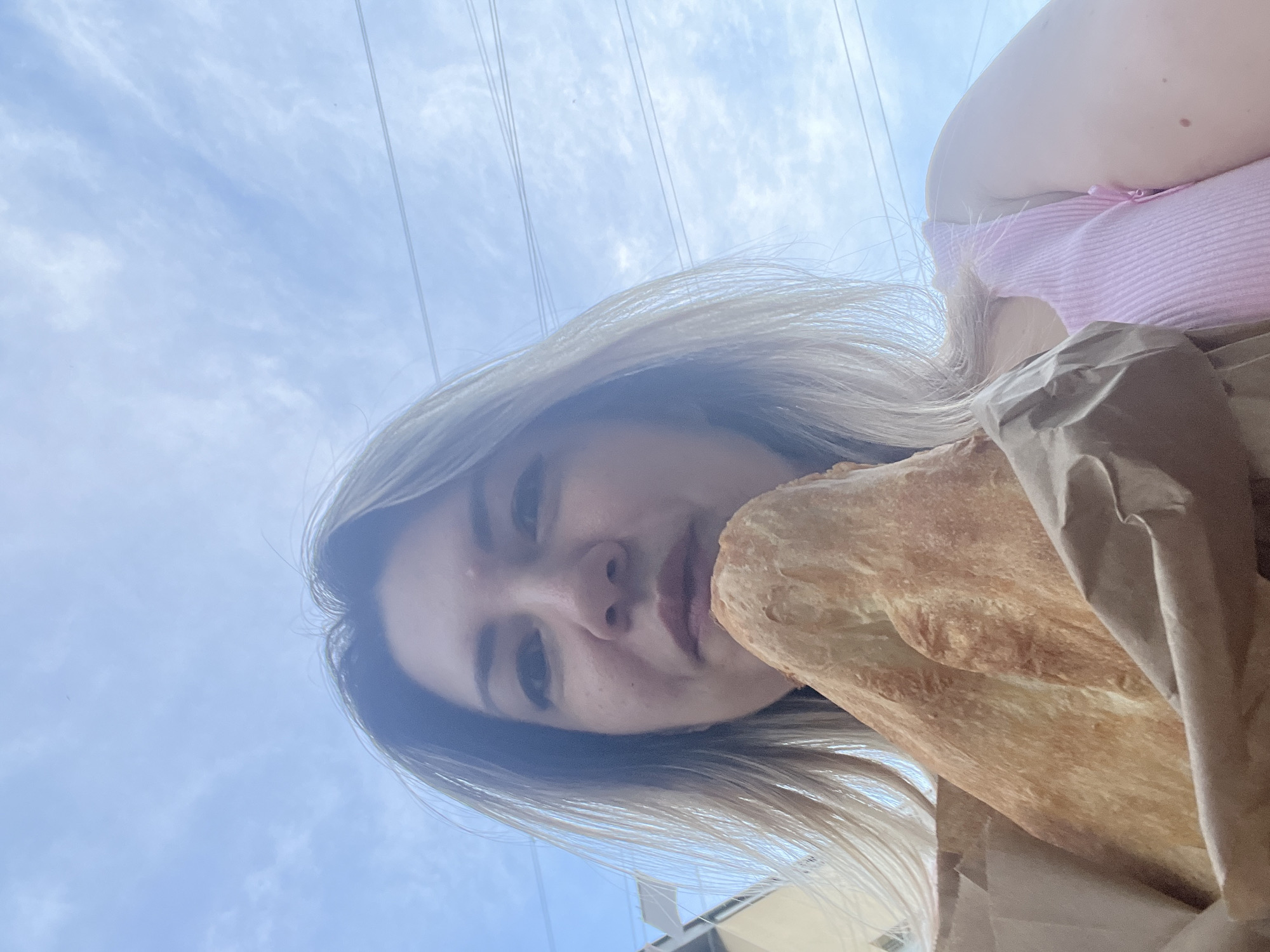 A woman with blonde hair, wearing a pink tank top, holds a large piece of bread wrapped in brown paper. She is looking at the camera, and her face is partially obscured by the bread. The background is a blue sky with scattered clouds and overhead power lines. The scene appears to be outdoors, possibly in the city of Sochi, Russia. The woman is holding the bread in front of her mouth as if about to take a bite. The lighting suggests it is daytime.