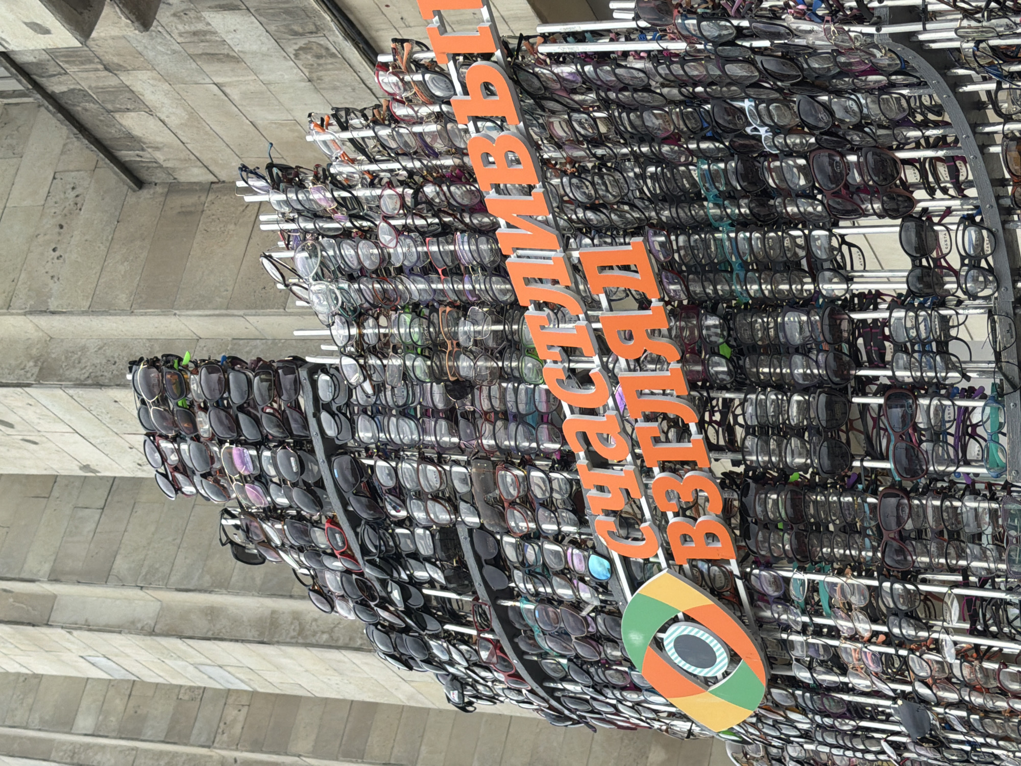 The image shows an outdoor display of sunglasses and eyeglasses in front of a building with a stone facade. The display is a tall, tiered structure filled with many pairs of eyewear, predominantly sunglasses. The eyewear is organized on metal racks. Above the eyewear, large orange letters spell out text in Russian. To the left of the text, there is a logo that resembles an eye within a colorful, segmented circle. The building facade appears to be made of large, rectangular stone blocks, consistent with classical or brutalist architecture. The sky is overcast. The location is identified as Saint Petersburg, Russia.