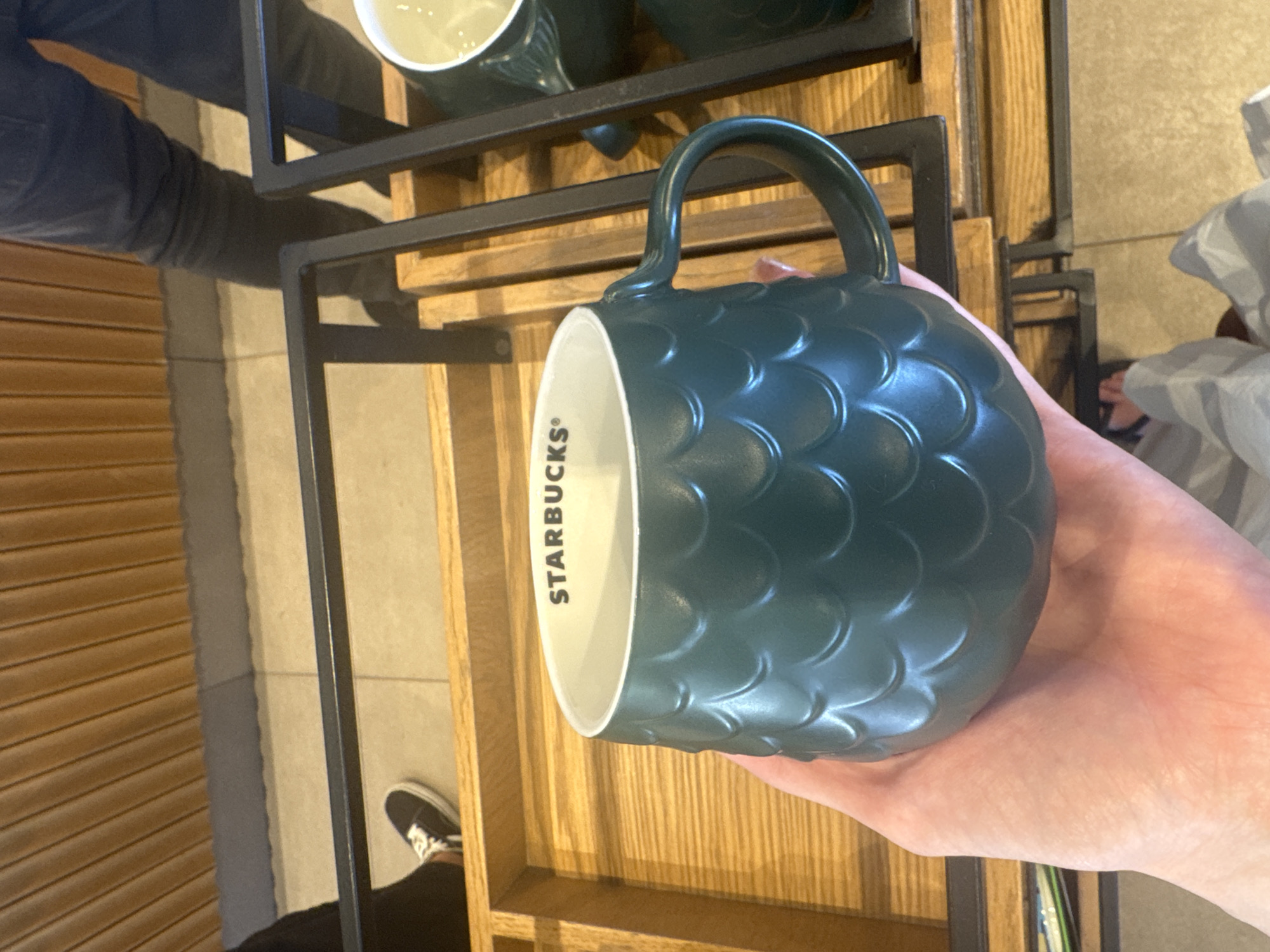 A hand holds a dark teal mug with a textured scale pattern. The mug has a white interior with the black Starbucks logo visible. The background shows wooden slats and black metal shelving, suggesting a retail environment. To the left, a person's lower legs and feet wearing black sneakers with white laces are visible. To the right, another dark teal mug is partially visible on a shelf, containing a light-colored liquid. The floor appears to be tiled. The scene is indoors, likely a Starbucks store.