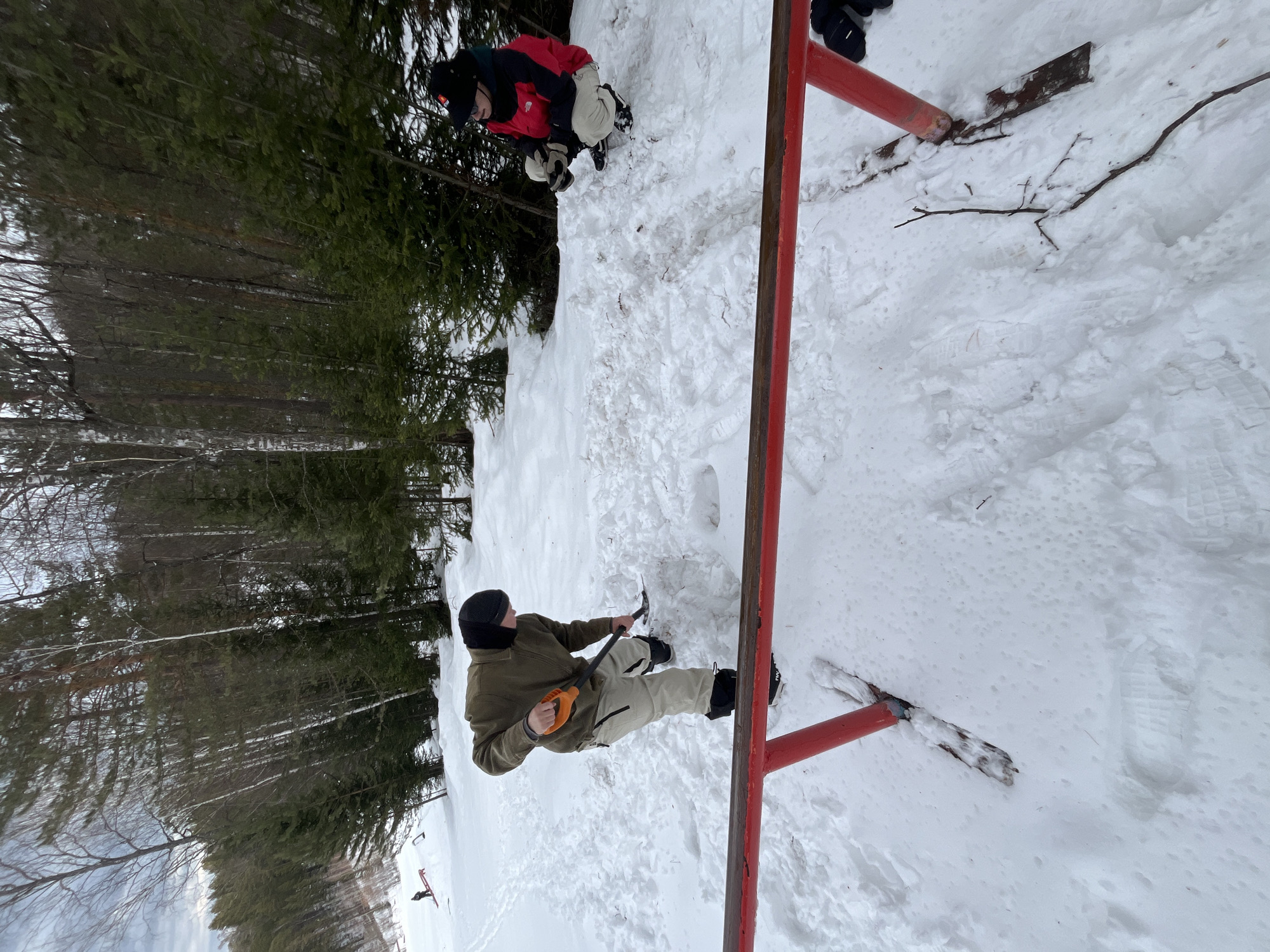 The image depicts a snowy outdoor scene with two individuals present. One individual is standing in the snow, wearing a black beanie, a green jacket, and tan pants, holding a tool that appears to be a pickaxe. The second individual is further away, also dressed for cold weather with a black beanie and a red and black jacket, and appears to be kneeling or crouching in the snow. A red metal structure is prominently in the foreground, adding to the scene's depth. The background features a forest with trees covered in snow, suggesting a winter environment. The location is Unknown, Unknown.