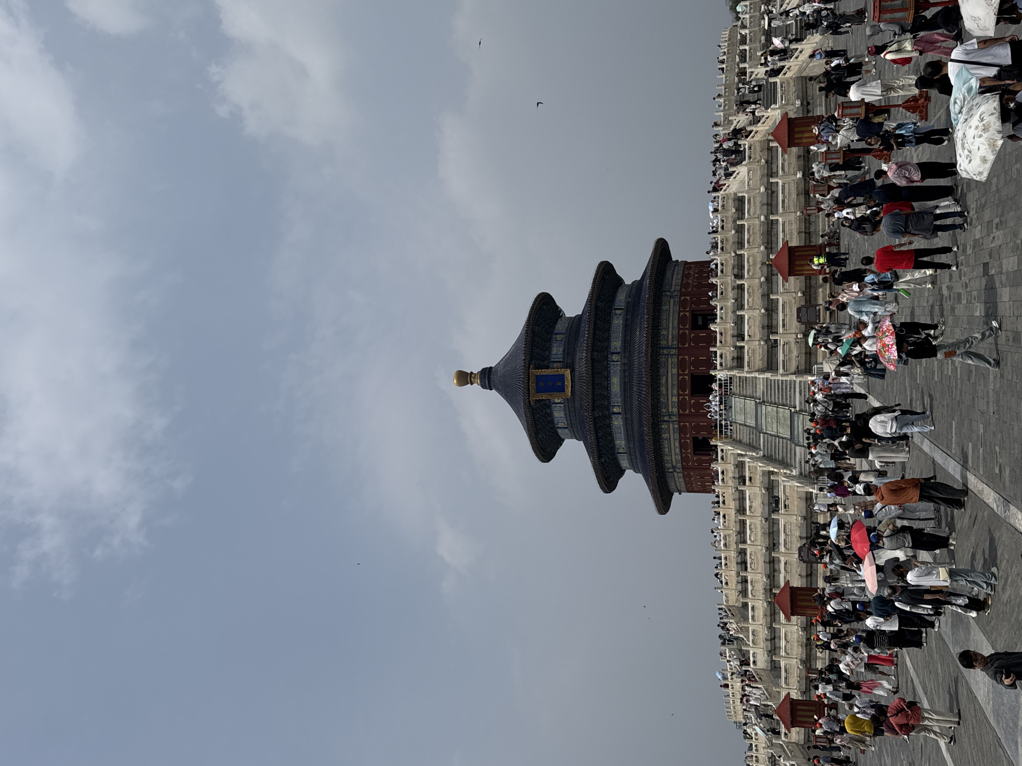 A vertical eye-level outdoor shot depicts the Temple of Heaven, situated in the Dongcheng District of Beijing, China. The Hall of Prayer for Good Harvests, a prominent feature of the temple, is seen with its tiered, dark-colored roof and gold-colored finial. A multitude of people are visible on the stone plaza surrounding the temple, creating a dense gathering. The sky overhead is light blue-gray with scattered clouds. Several birds are seen flying in the sky.