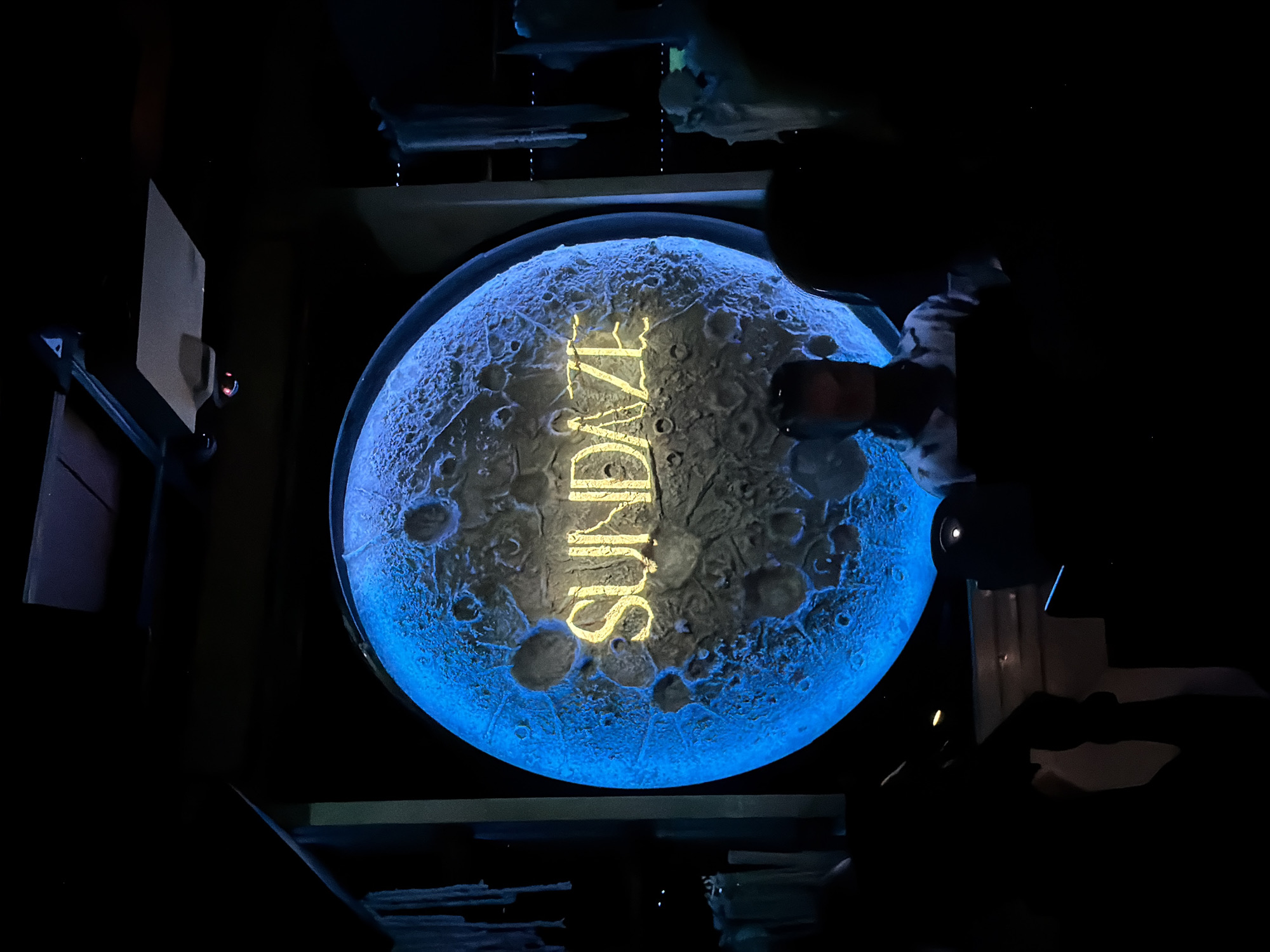 The image shows a dimly lit indoor space with a large, circular projection of the moon or a similar celestial body, illuminated by a bright blue light. The word "SUND AZE" is prominently displayed in yellow, block lettering across the surface of the projection, which is textured to resemble craters. In the foreground, silhouettes of several people are visible, facing the projection. The environment appears to be a public space, possibly an entertainment venue or a display in a city like North Kuta, Indonesia, given the mention of its location. The overall atmosphere is dark and somewhat theatrical, with the bright projection serving as the central focal point.