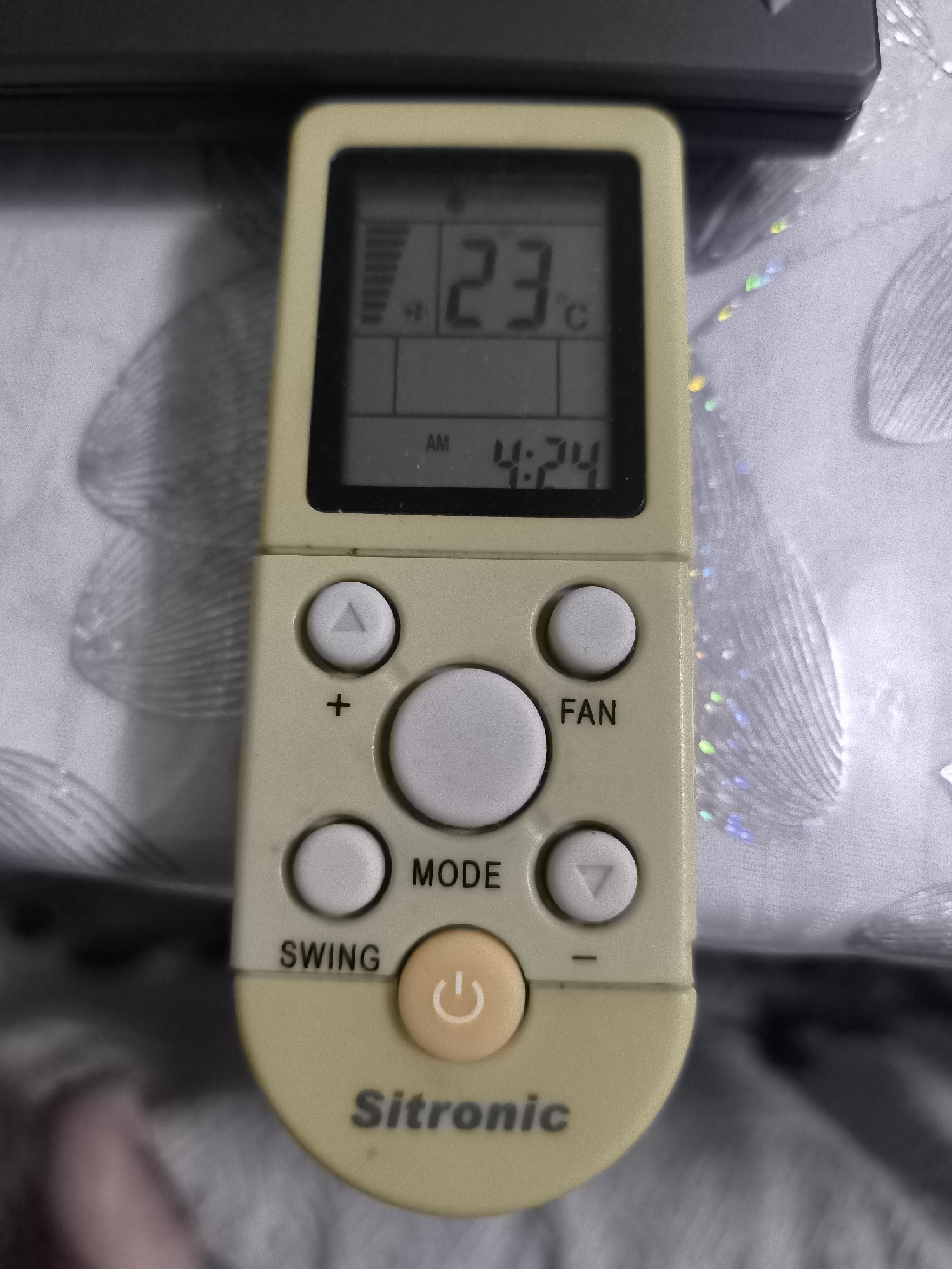 This is a close-up, eye-level shot of a beige air conditioner remote control with a digital display. The remote is positioned vertically in the center of the frame, resting on a textured, light-colored surface that appears to be fabric, possibly a tablecloth or sofa upholstery, with a patterned design. The background is blurred, showing more of the same fabric and dark, indistinct shapes above.
The digital display on the remote shows "23°C" at the top, indicating the current temperature. Below that, it displays "AM 4:24", suggesting the time. The remote has several buttons: a circular power button with a white circular icon, a larger central button labeled "MODE", and other smaller buttons arranged around it, including directional arrows (up and down triangles), a plus sign, and a button labeled "FAN". There's also a button labeled "SWING". The brand name "Sitronic" is visible in dark text at the bottom of the remote. The overall lighting is soft, and there are no explicit indications of weather or specific emotions. The context of Kokand, Uzbekistan, is provided but not visually discernible from the image itself. The focus is entirely on the remote control.