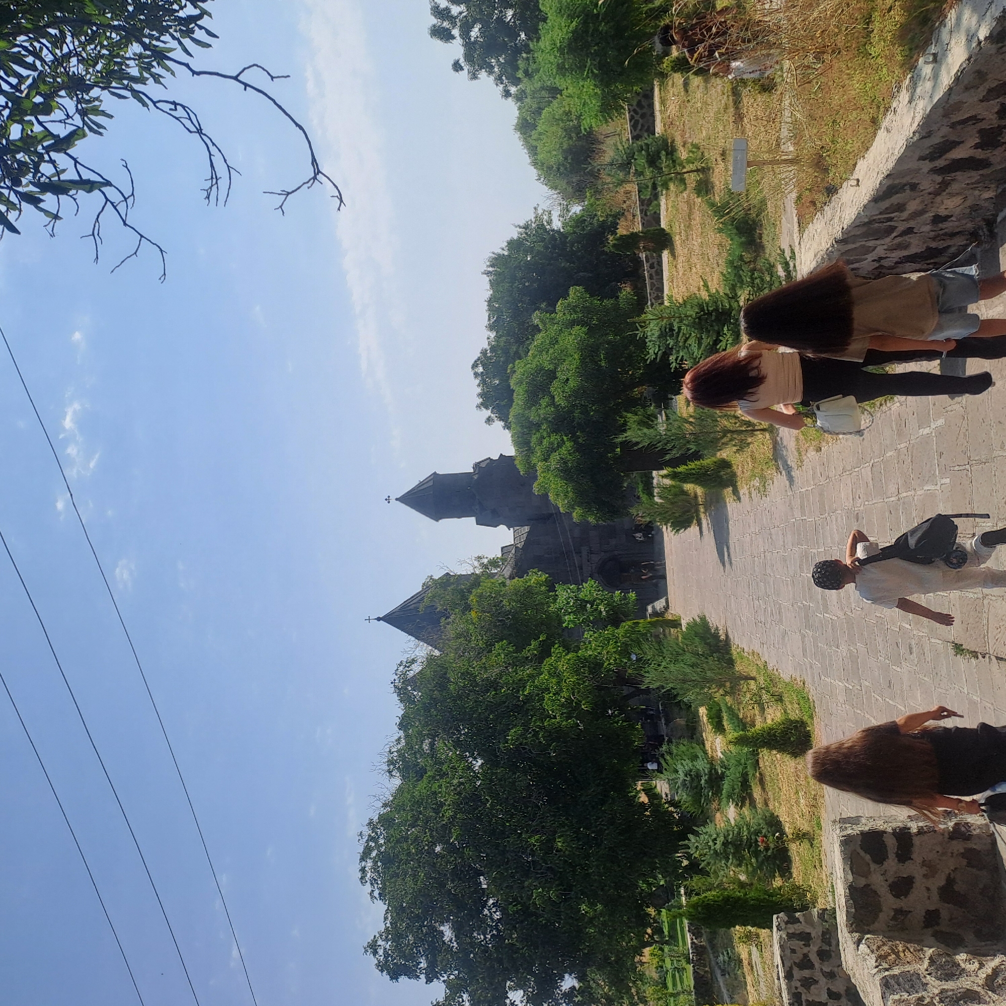 In Tegher, Armenia, a group of people are walking up a stone pathway towards a church. The pathway is lined with stone walls and manicured bushes. A large, old church with two prominent towers and crosses on top is visible in the background, surrounded by lush green trees. The sky is a clear blue with a few wispy clouds and three thin wires stretched across it. The people, including a woman in a beige top and black pants carrying a white bag, a young boy in a white outfit with a black backpack, and two girls with long hair, are all walking away from the camera up the path. The environment is a well-maintained park or historical site.
