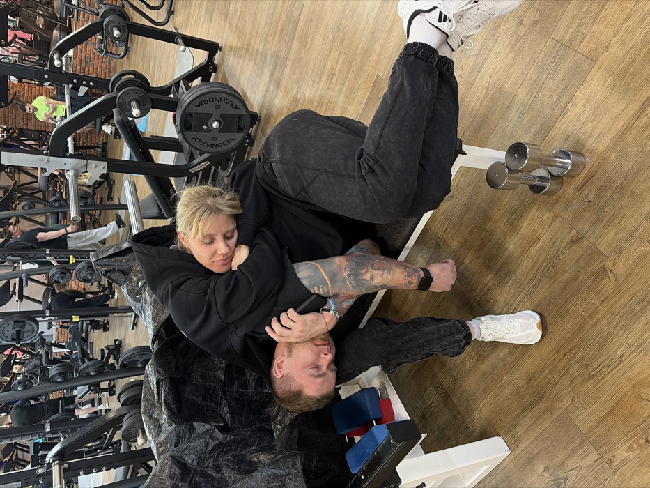 In a gym located in Chaikovsky, Russia, two individuals are depicted resting on a white exercise bench. An adult female, with blonde hair, is lying with her eyes closed, her head resting on the shoulder/chest area of an adult male. She is wearing a dark hooded sweatshirt and dark sweatpants, with white socks and white and black athletic shoes. The adult male, with short brown hair and light stubble, is also lying with his eyes closed, his head against the backrest of the bench. He is wearing a dark hooded sweatshirt and dark, stone-washed sweatpants, with white athletic shoes. His right arm, adorned with a full-sleeve tattoo featuring a portrait of a person and other designs, has a black wristband. A dark, crumpled tarp-like material covers part of the bench to the left of the male. Two chrome dumbbells are positioned on the light wood-grain floor in front of the bench. In the background, numerous other individuals are visible, some actively using various gym equipment including weight machines, benches, and barbells. Exposed brickwork and painted walls are visible behind the equipment.