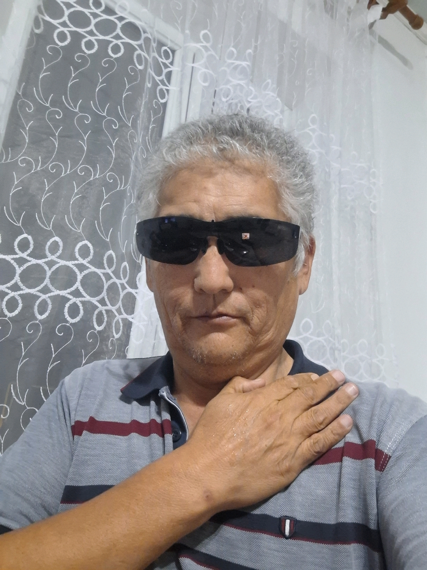 A middle-aged man with gray hair and sunglasses is taking a selfie in front of a window with white lace curtains. He is wearing a gray polo shirt with dark blue and maroon stripes. His right hand is placed on his chest. The background appears to be an indoor setting, likely a home. No specific location within Almaty, Kazakhstan can be determined from the image.