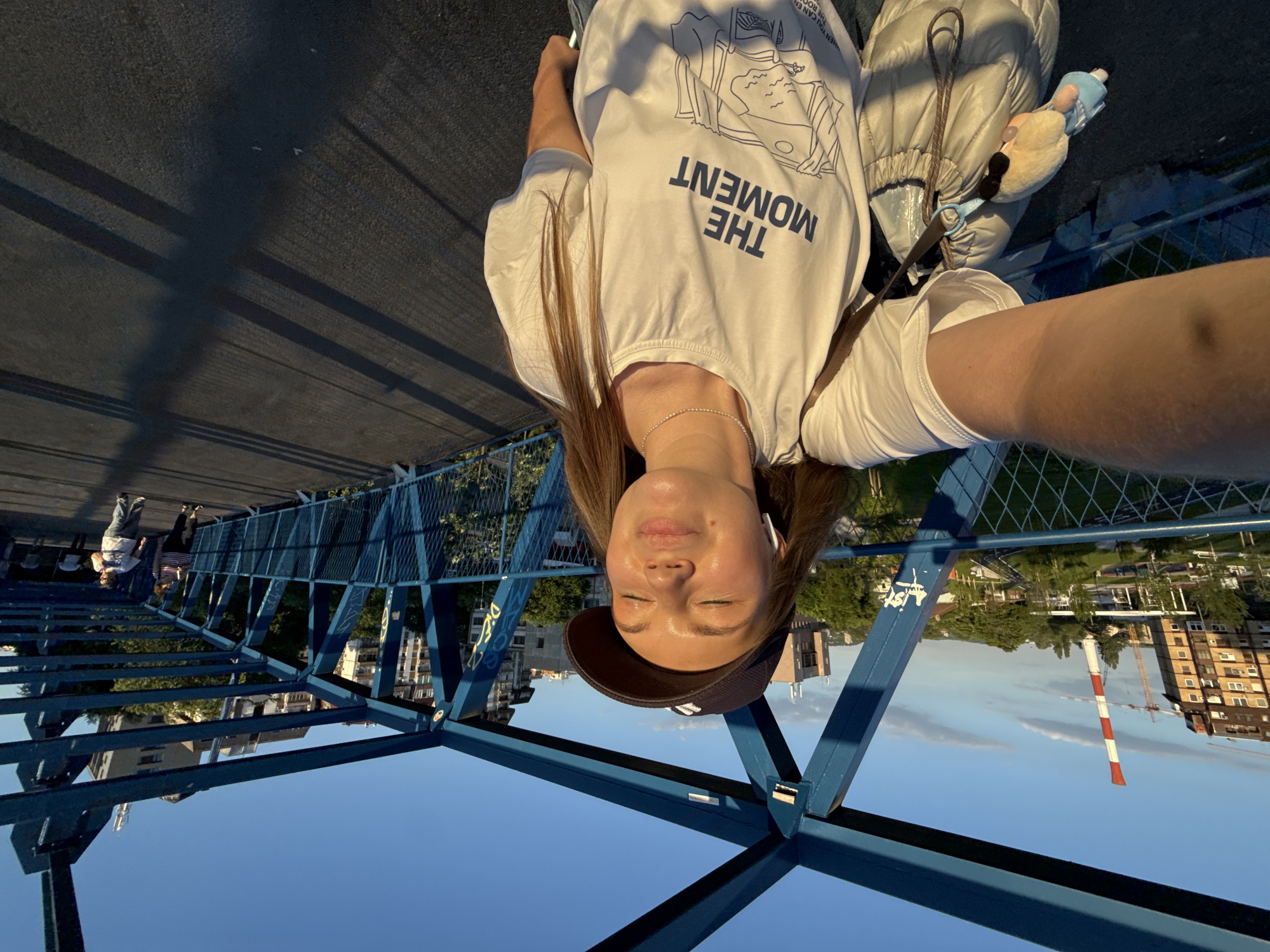 A selfie taken by a woman on a blue-painted steel pedestrian bridge. The woman is wearing a white T-shirt with "THE MOMENT" printed on it, a necklace, a dark cap, and earbuds. She is facing the camera with her eyes closed. Attached to her jacket is a small plush toy. In the background, urban buildings and a red-and-white chimney can be seen. Other pedestrians are walking on the bridge in the distance. The location is likely Belgrade, Serbia, based on the architectural style and the presence of a chimney.