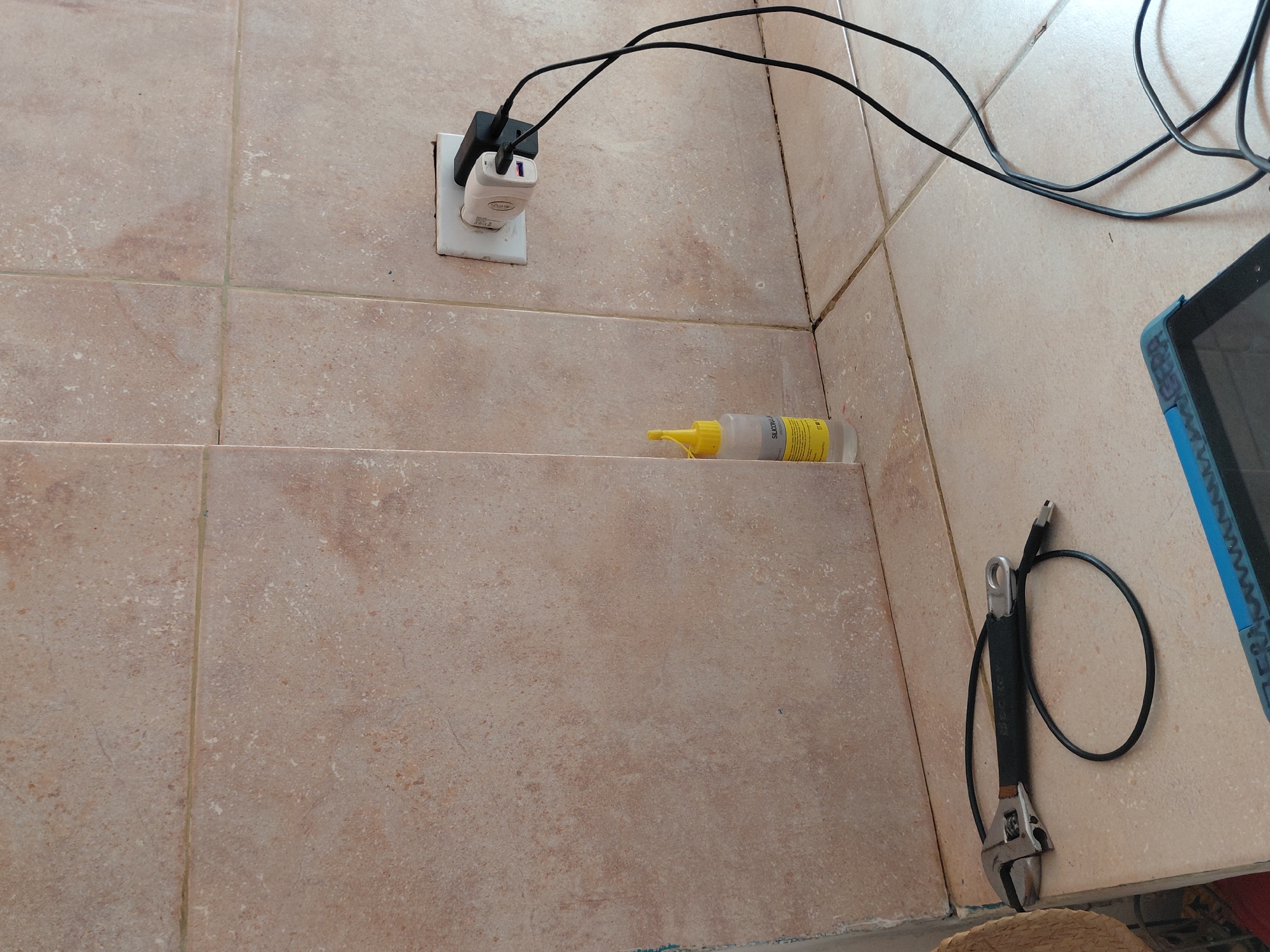 The image captures a corner of an indoor space, likely in Santa Ana, Venezuela, characterized by light brown/beige tiled walls and surfaces with visible grout lines. An electrical outlet is installed on the upper tiled wall, into which two white USB wall chargers are plugged. One charger has a black adapter also plugged into it. Multiple black cables extend from these chargers, disappearing out of frame to the right, suggesting devices are being charged.
On a lower tiled surface, to the right of the outlet, sits a translucent bottle with a yellow pointed cap. The bottle is labeled "SILICON" and "30g", and further text indicates it is an "ADHESIVO SELLADOR ELÁSTICO" (elastic adhesive sealant), "INCOLORO" (colorless), and for "USO MÚLTIPLE," suitable for various materials like glass, aluminum, ceramic, plastics, wood, metals, and stone.
To the far right, on another tiled surface, an adjustable wrench with a black handle grip is visible. A black cable, possibly another charging cable, is wrapped around the wrench. Partially visible in the top right corner is a blue-cased electronic device, resembling a tablet or small laptop, with the word "RESERVA" and other indistinct text printed on its blue cover. The overall impression is a functional, somewhat utilitarian space where items are casually stored or used. There are no people present, and no specific activity is actively depicted, other than the implied charging of devices.