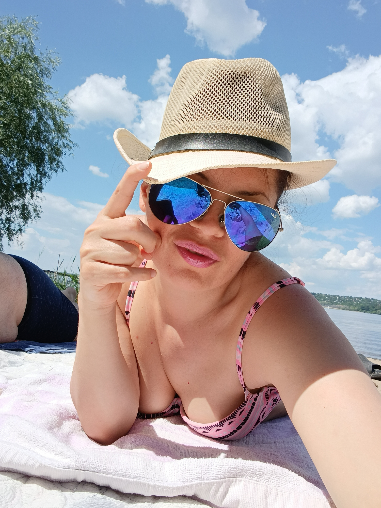 A woman wearing a straw hat and mirrored aviator sunglasses is lying on a pink and white striped bikini top on a towel. She is on a beach with a body of water in the background and trees and clouds in the sky. Another person's legs wearing dark blue swim trunks are visible to the left of the woman. The scene is set on a sunny day.