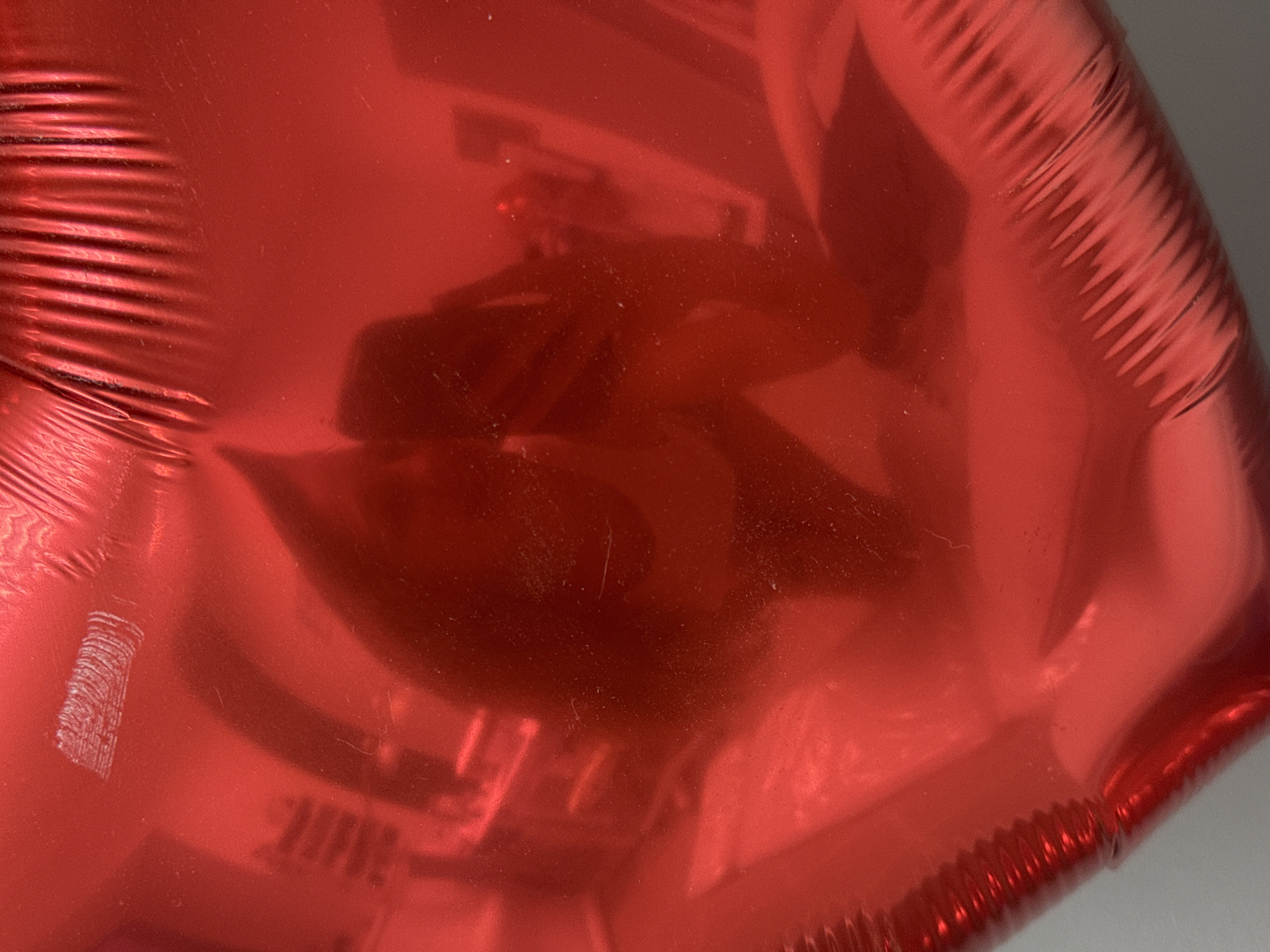 A close-up image shows a red, inflated, and crinkled foil balloon. The shiny surface reflects the surrounding environment, obscuring details of what the reflection is of. The color and shape suggest a festive or celebratory object. The background is a neutral gray color. Izhevsk, Russia, cannot be determined from this picture.