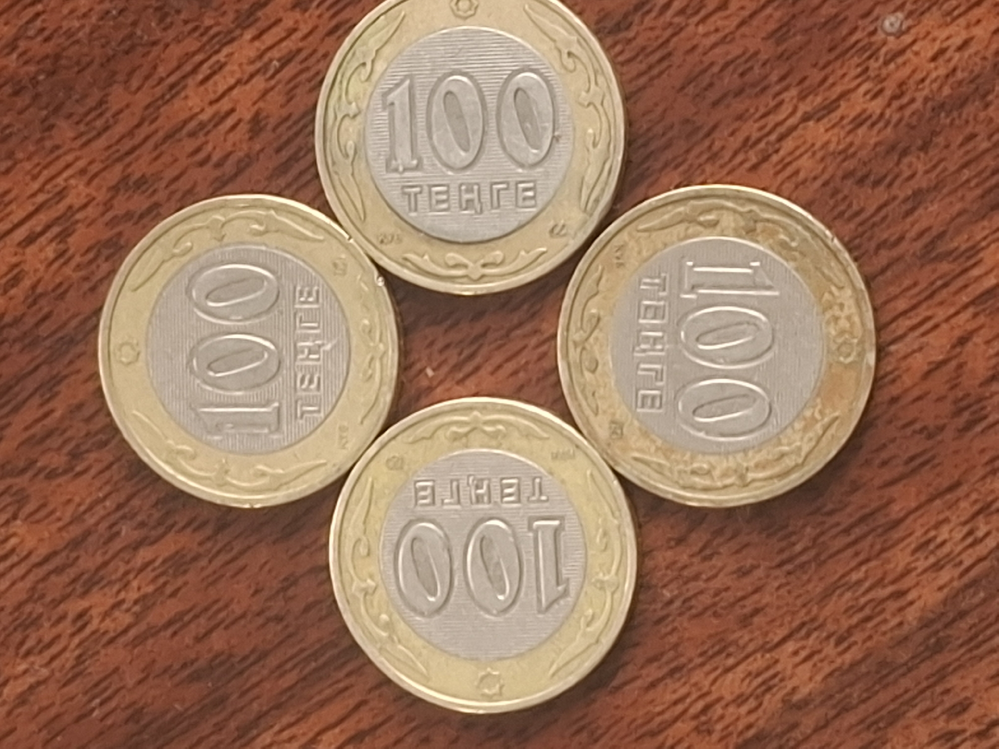 The image shows four coins, each with the number "100" and the word "TENGE" written on them. The coins are arranged in a diamond shape on a wooden surface. The wooden surface has a reddish-brown hue and a visible grain pattern, suggesting it might be a table or countertop. The lighting appears to be from above, casting subtle shadows from the coins. The coins themselves have a bi-metallic design, with a silver-colored center and a gold-colored outer rim. The image does not contain any people or depict any specific location in Almaty, Kazakhstan.