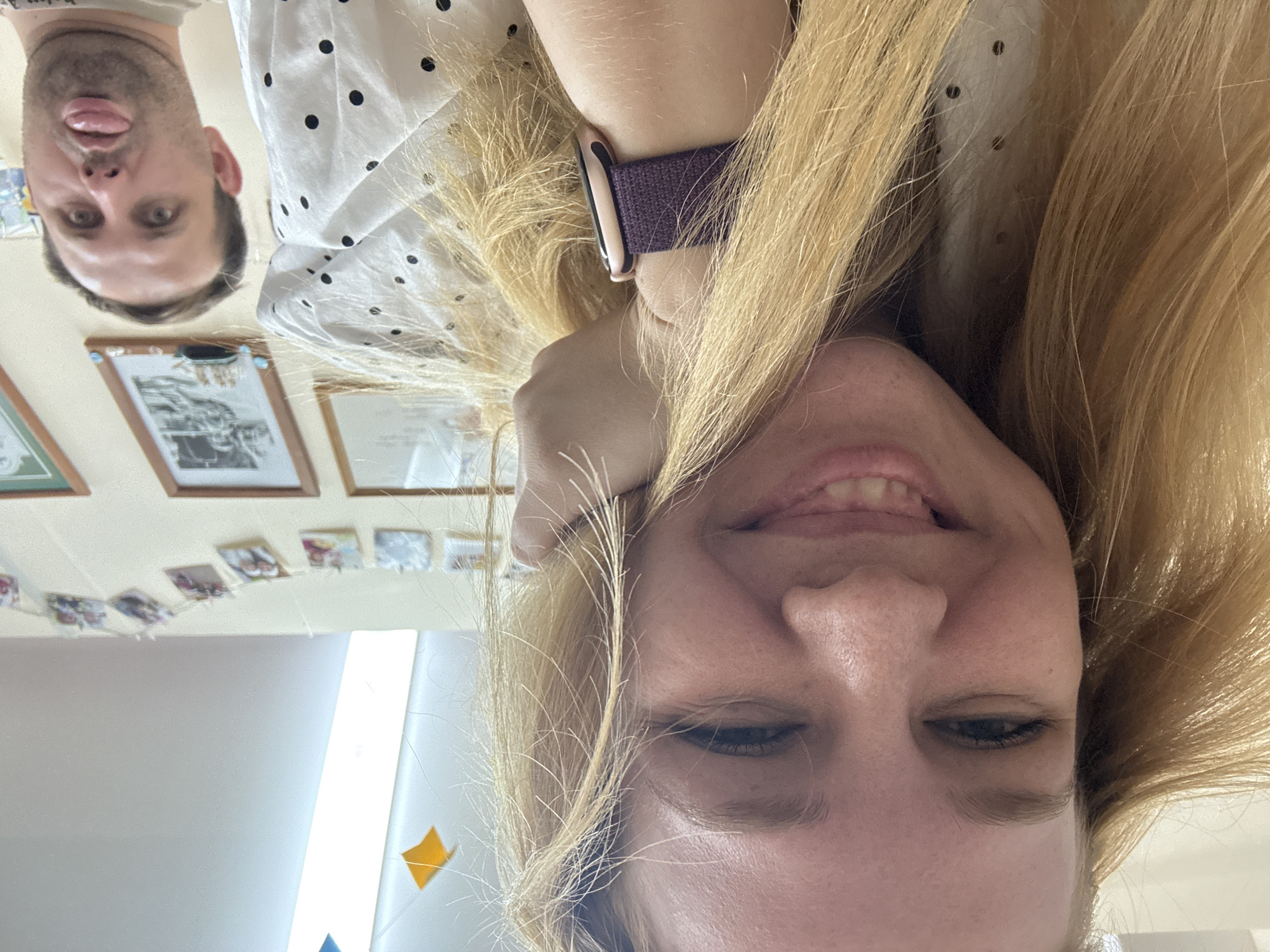 The image depicts two individuals. One is a fair-skinned male with short brown hair, sticking his tongue out. The second person is a fair-skinned female with long blonde hair, smiling at the camera while holding her hand to her face. She is wearing a white shirt with black polka dots and a purple Apple Watch band. The background includes a cream-colored wall adorned with framed photographs and a string of small, colorful images. The location is not discernible from the provided image. No specific details indicate the location is in Rusanovo, Russia.