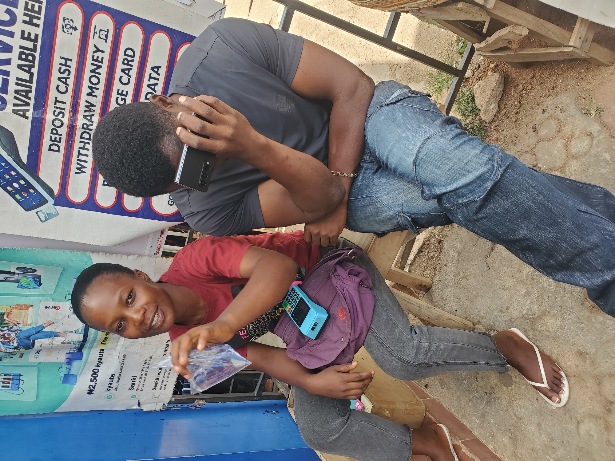The image depicts a man and a woman sitting outside a shop in Jos, Nigeria. The man, wearing a gray t-shirt and blue jeans, is sitting in the background, talking on a smartphone. The woman, sitting in the foreground, is wearing a red t-shirt and gray jeans. She holds a plastic bag in her hand and smiles at the camera. A point-of-sale terminal hangs from her waist. Behind them is a sign advertising deposit cash, withdraw money, recharge card, and data services. A poster of a television is also visible.
