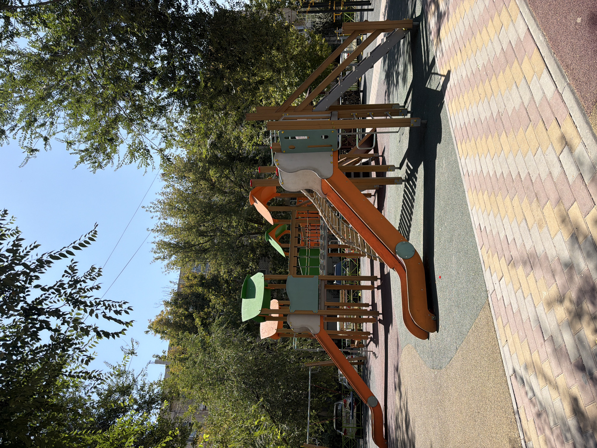 A modern children's playground is visible in the foreground, situated in a city environment, likely within Shymkent, Kazakhstan. The playground features an orange and grey slide, a rope bridge, and a climbing wall. The ground surface is a combination of rubberized safety surfacing and decorative brickwork. Dense green trees surround the playground, casting shadows on the ground. In the background, a multi-story building and parked cars are visible. The sky is clear and blue. There are no people or animals present in the image.